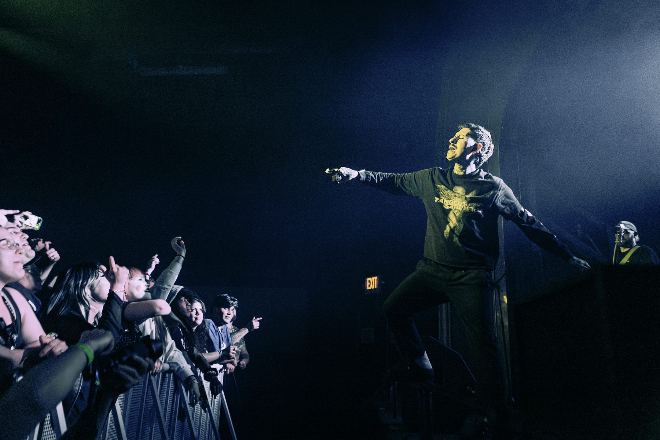 A live performance at a concert featuring a male singer on stage engaging with the audience, with fans reaching out and cheering in the foreground. The venue is dimly lit, highlighting the performer.