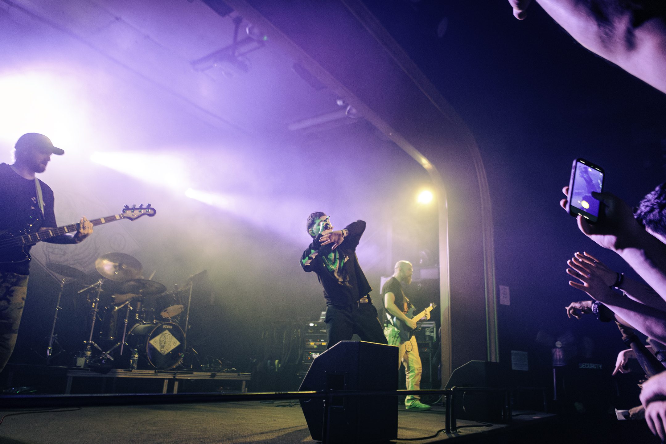 A live performance scene featuring a musician in a ski mask with hands raised, flanked by band members playing guitar and drums, under vibrant stage lights.