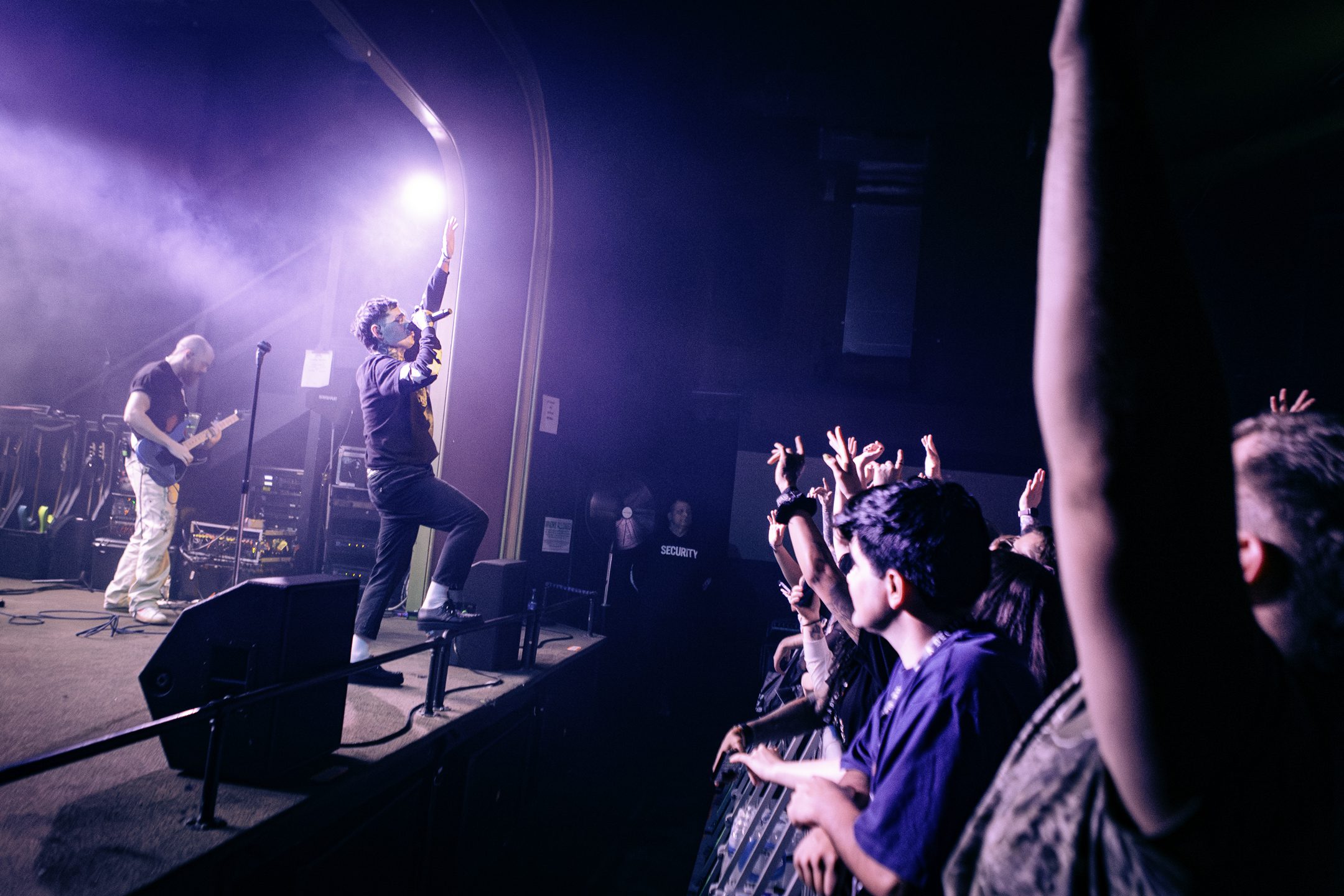 A live performance featuring a singer holding a microphone and passionately engaging with the audience, while a guitarist plays in the background. The crowd eagerly raises their hands, illuminated by purple stage lighting.