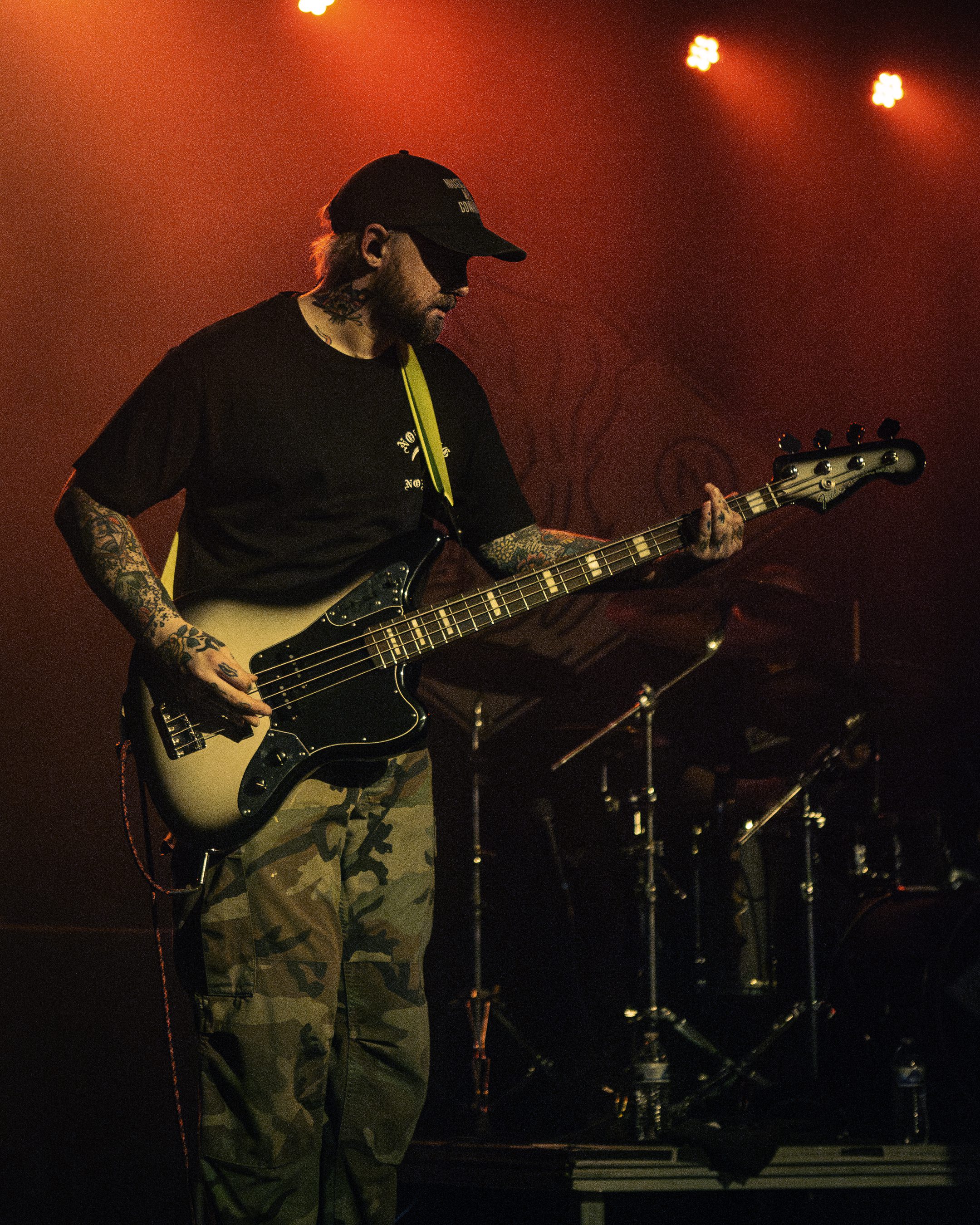 A musician playing a bass guitar on stage, dressed in a black t-shirt and camouflage pants, with tattoos visible on their arms. The stage is lit with red lighting, creating a dramatic atmosphere.
