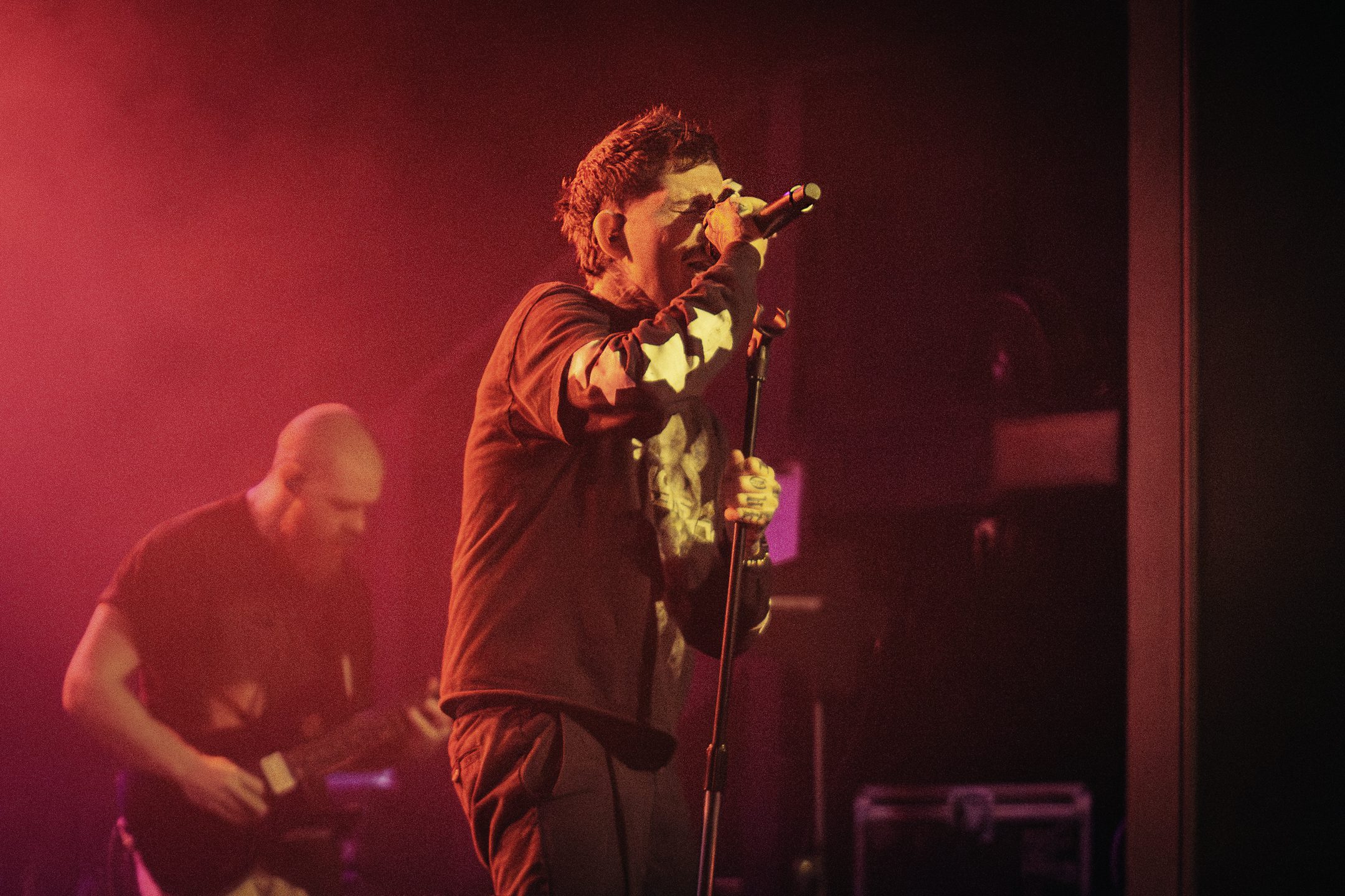 A male musician performing on stage with a microphone, under pink lighting, while another musician plays guitar in the background.