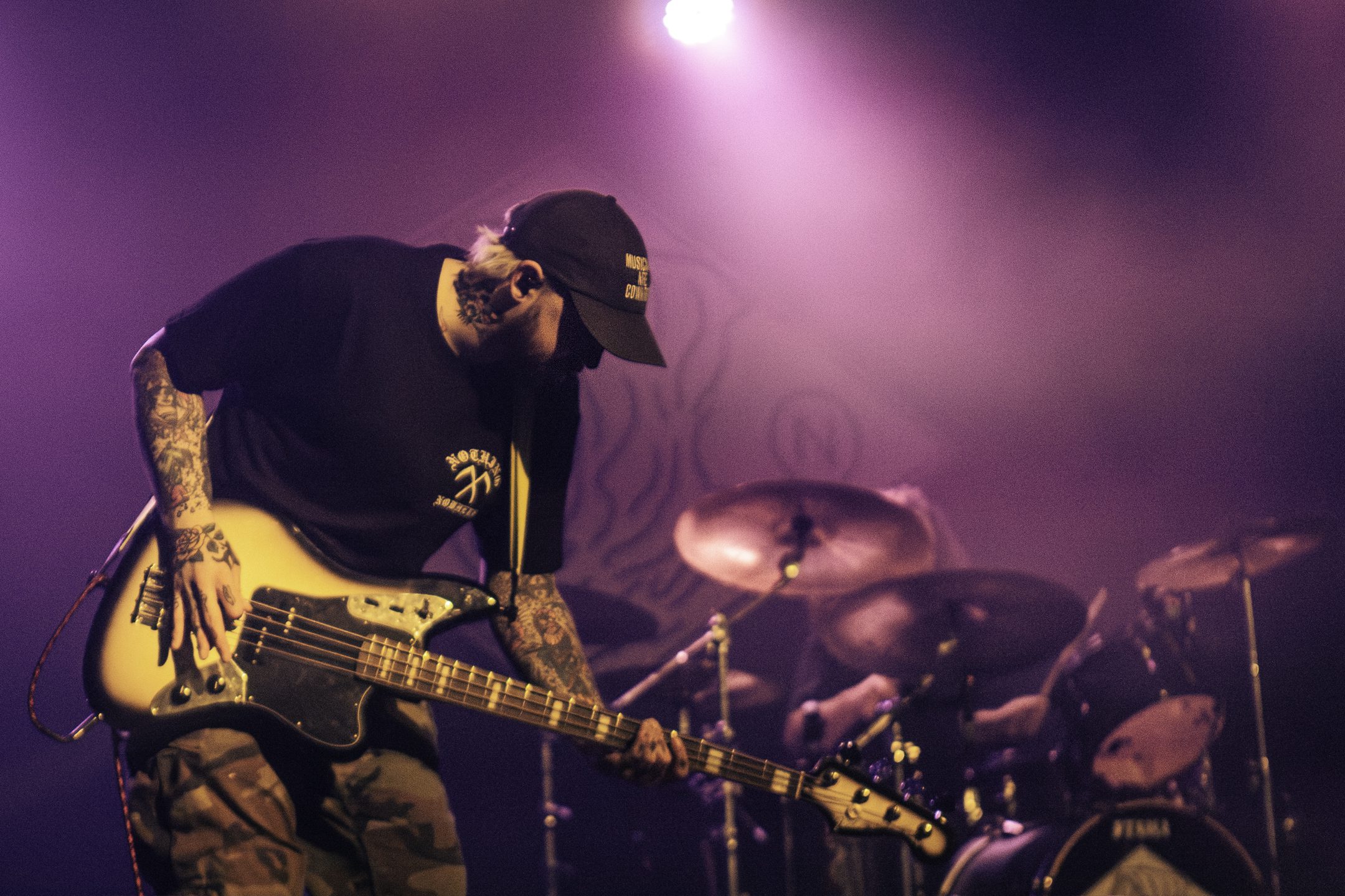 A musician playing a bass guitar on stage with a tattooed arm, wearing a black shirt and cap, with drums visible in the background under purple lighting.