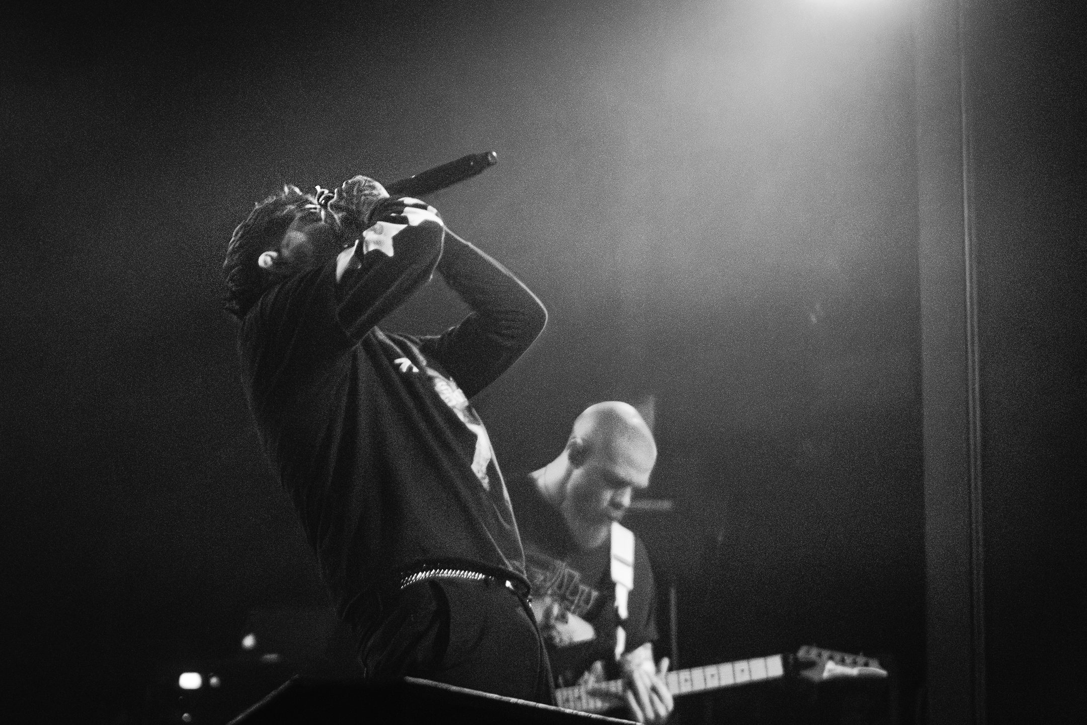 A male musician performing on stage with a microphone, showing intense emotion, while a guitarist plays in the background, illuminated by stage lights.