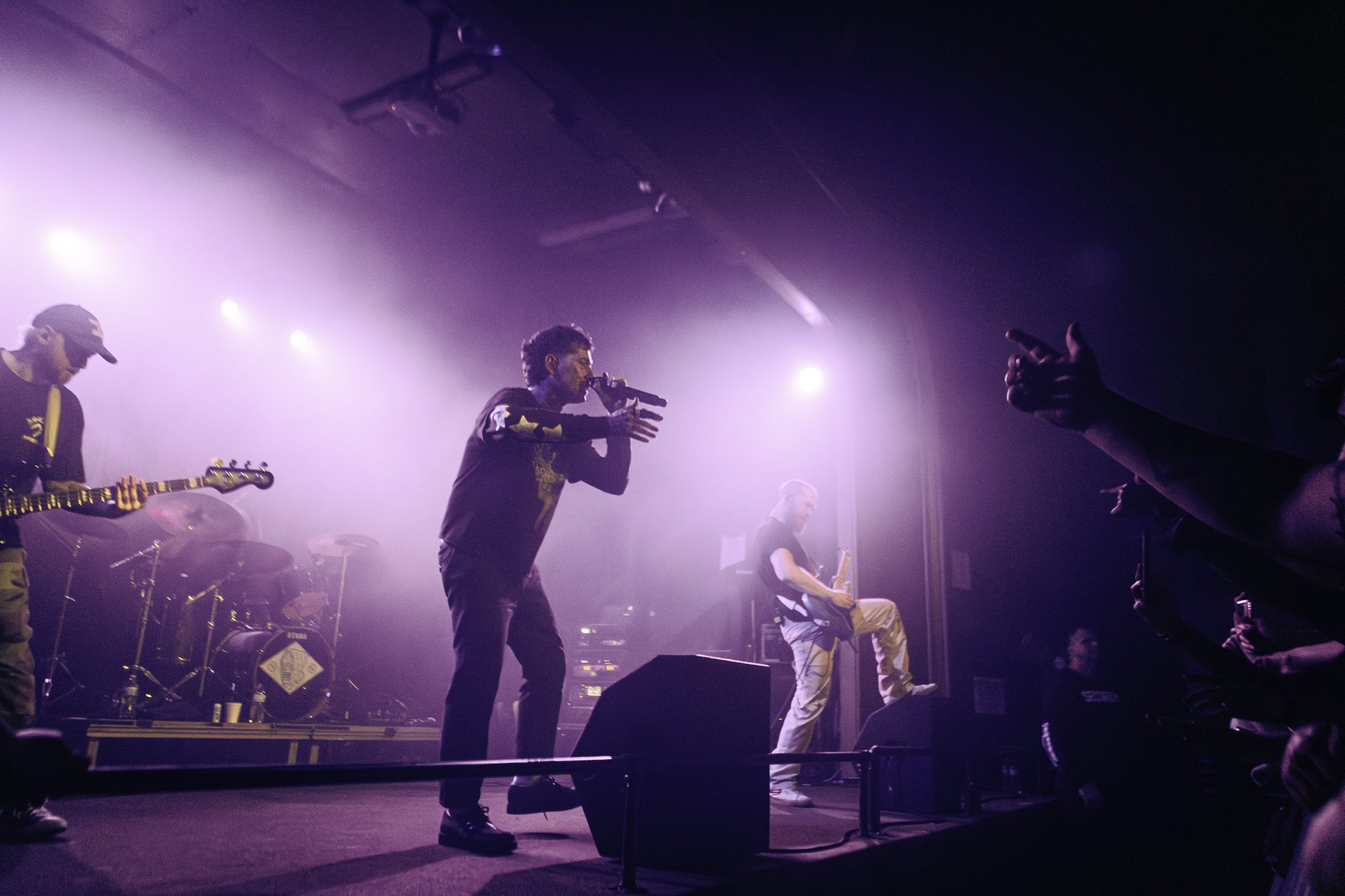 A lively concert scene featuring a vocalist passionately performing on stage with a microphone, surrounded by a band playing instruments. The crowd is engaged, with hands raised in excitement, illuminated by purple stage lighting.