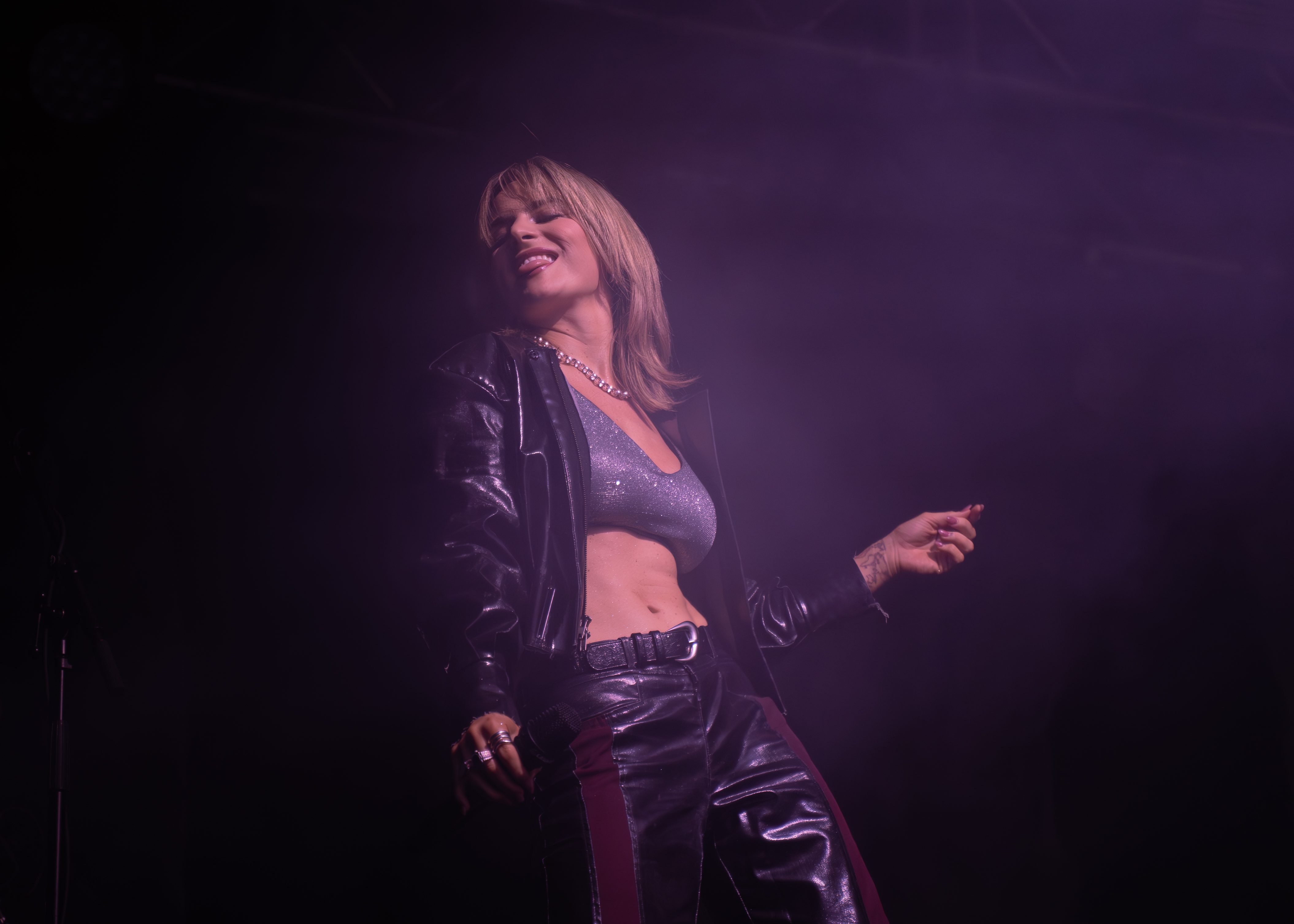 A performer with short blonde hair wearing a sparkling gray top and black leather jacket, striking a dynamic pose on stage with purple lighting in the background.