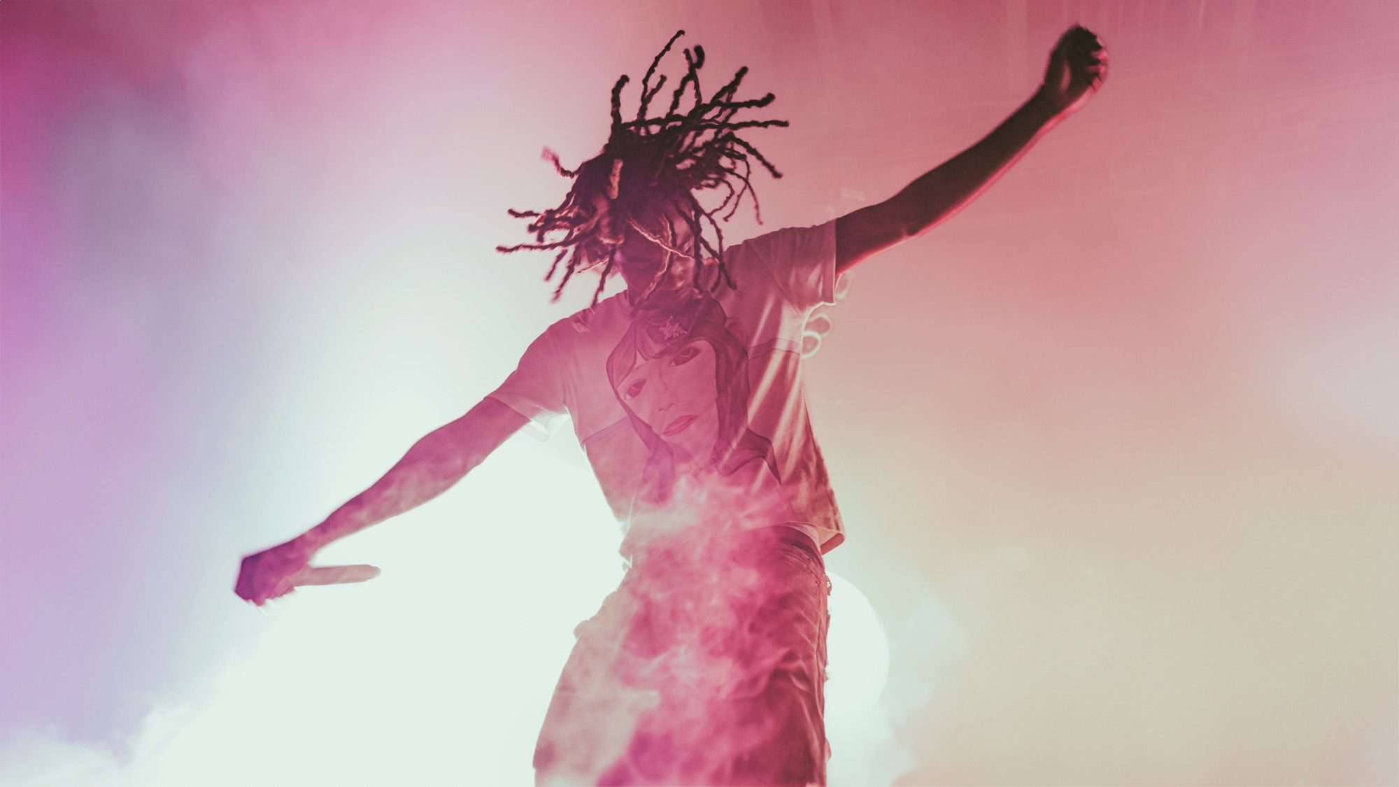 A performer with dreadlocks is energetically dancing on stage under colorful lighting, creating an electric atmosphere during a concert.
