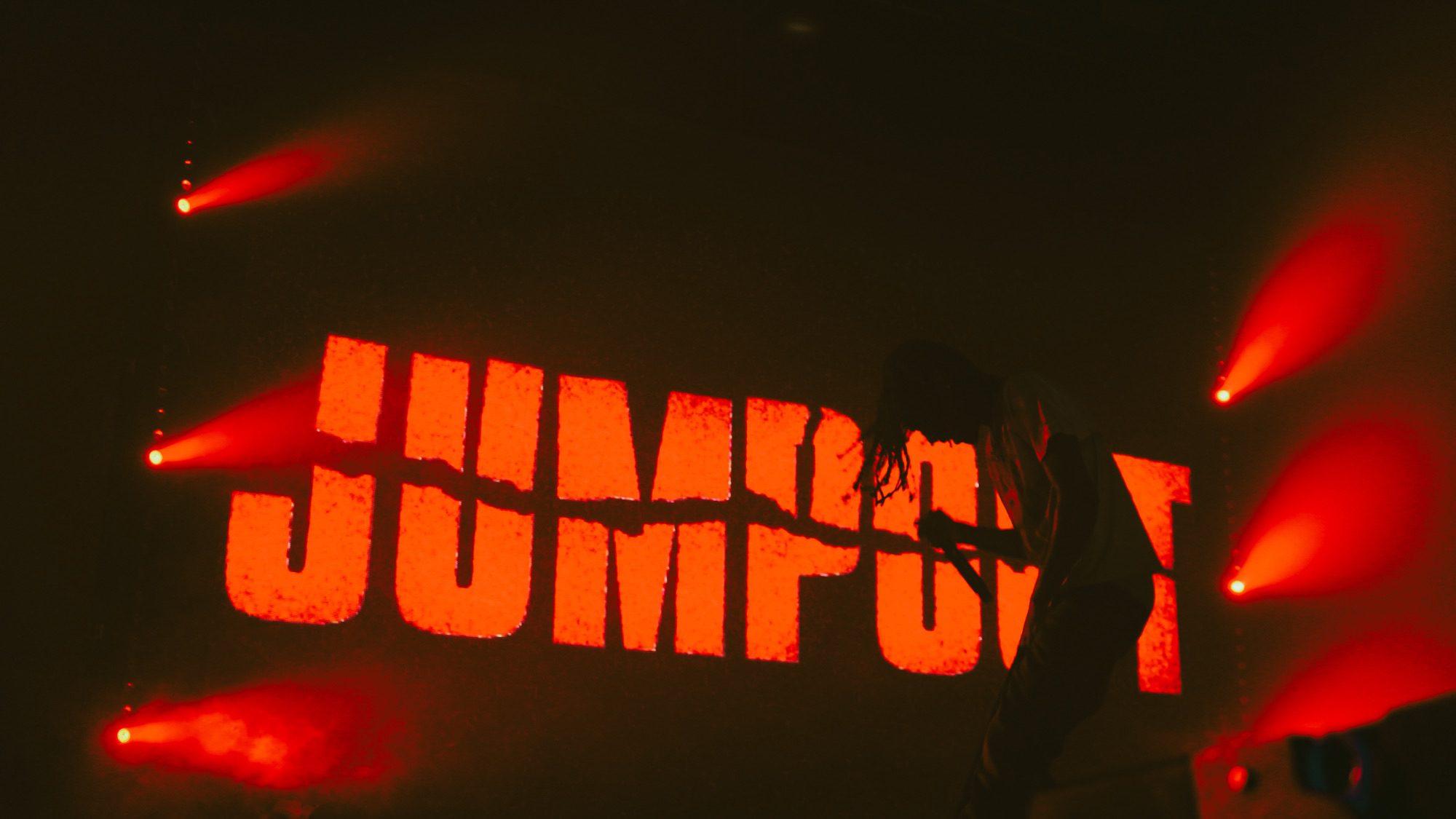 A silhouette of an artist performing on stage against a vibrant red backdrop featuring the word 'JUMPOUT' illuminated in bold letters, with stage lights creating an energetic atmosphere.