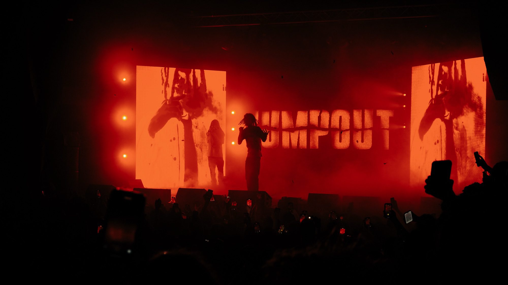 OsamaSon performing on stage during the JumpOut Tour, with vibrant red lighting and visuals behind him, while the crowd holds up their phones.