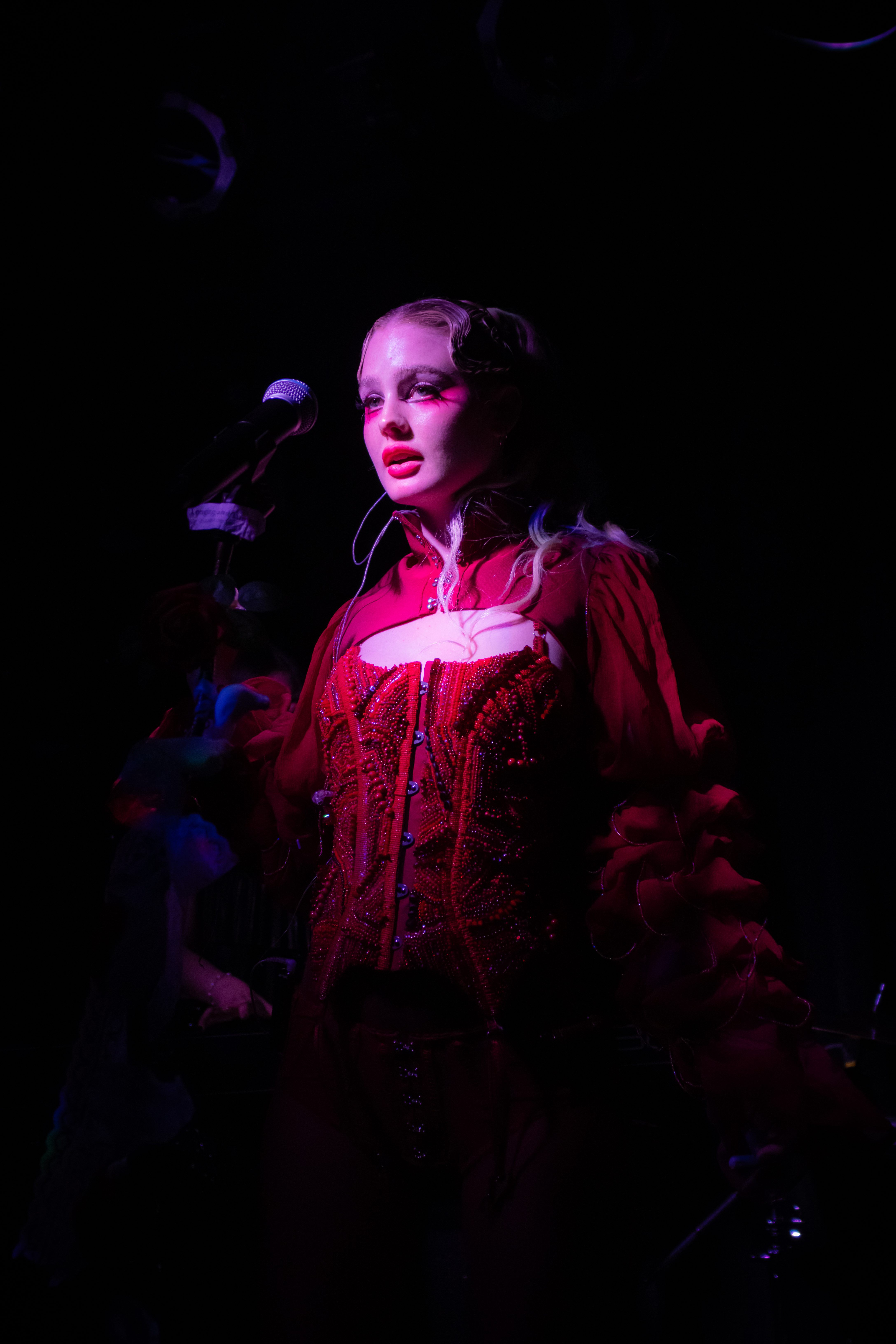A performer in a red beaded outfit stands on stage holding a microphone, illuminated by spotlight amid a dark background.