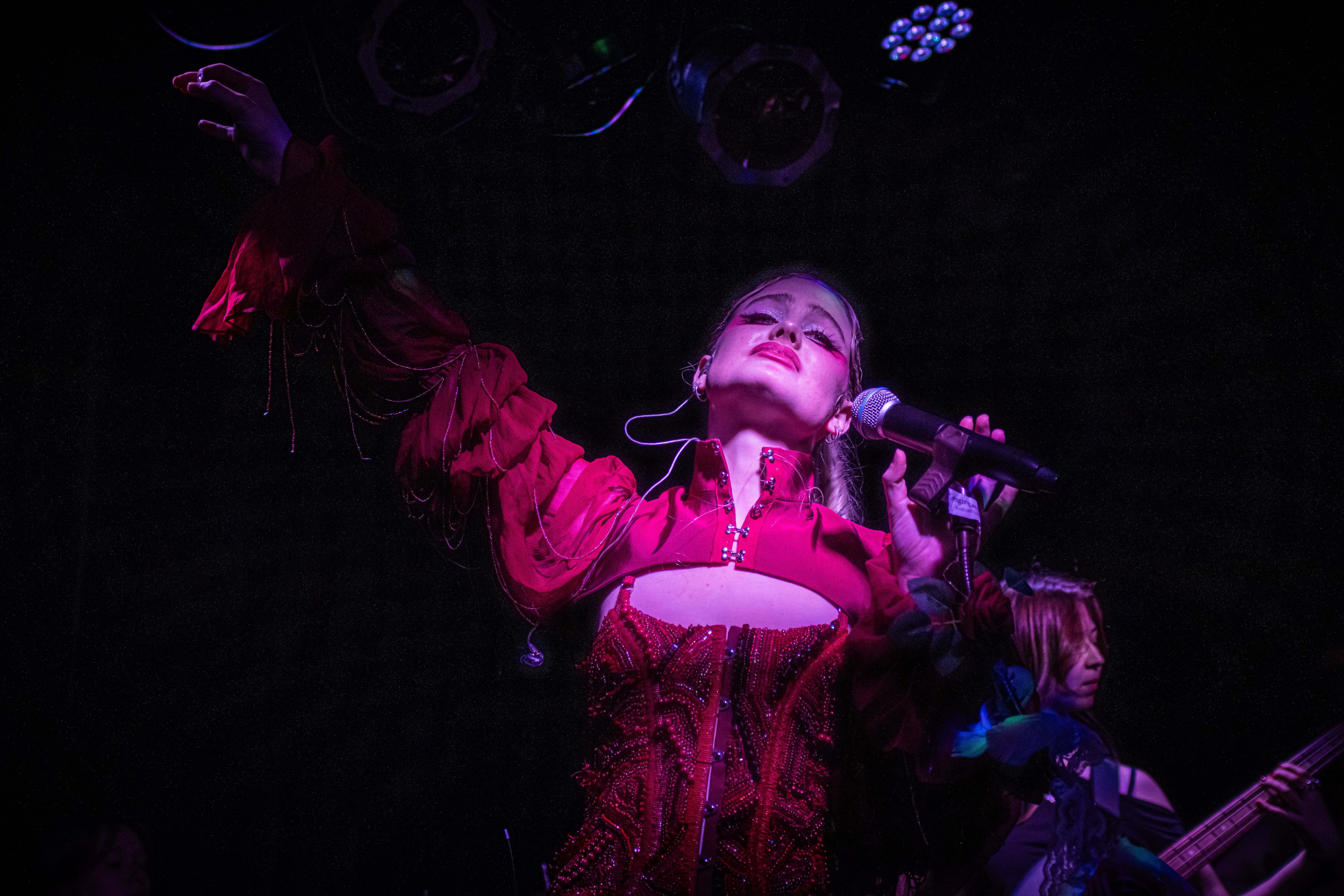 A female artist performing on stage, passionately singing into a microphone while wearing a vibrant red outfit, with dramatic lighting highlighting her expression and gestures.