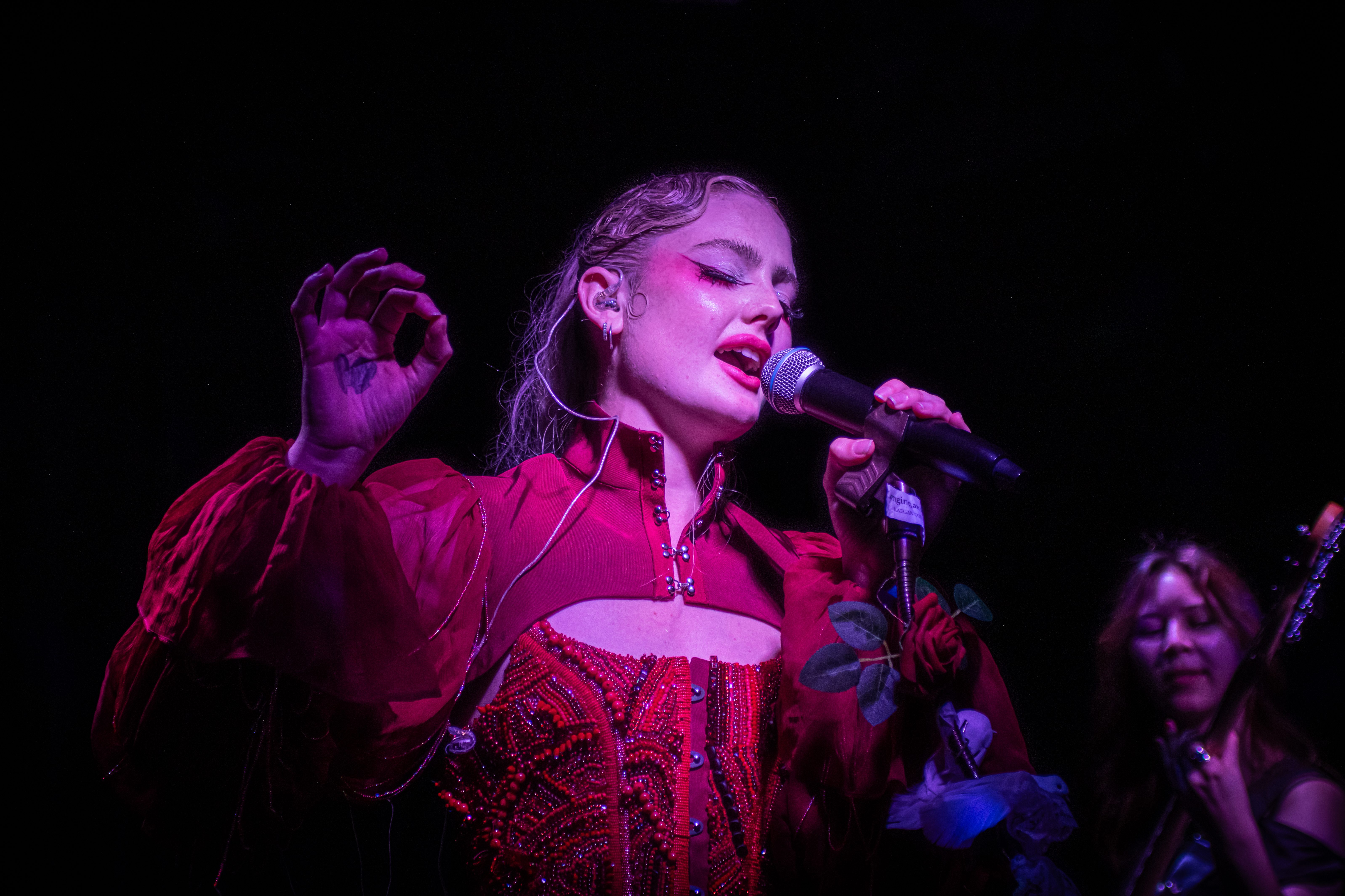 A female artist passionately performing on stage, holding a microphone and wearing an extravagant red outfit. A background vocalist can be seen behind her, contributing to the vibrant atmosphere of the performance.