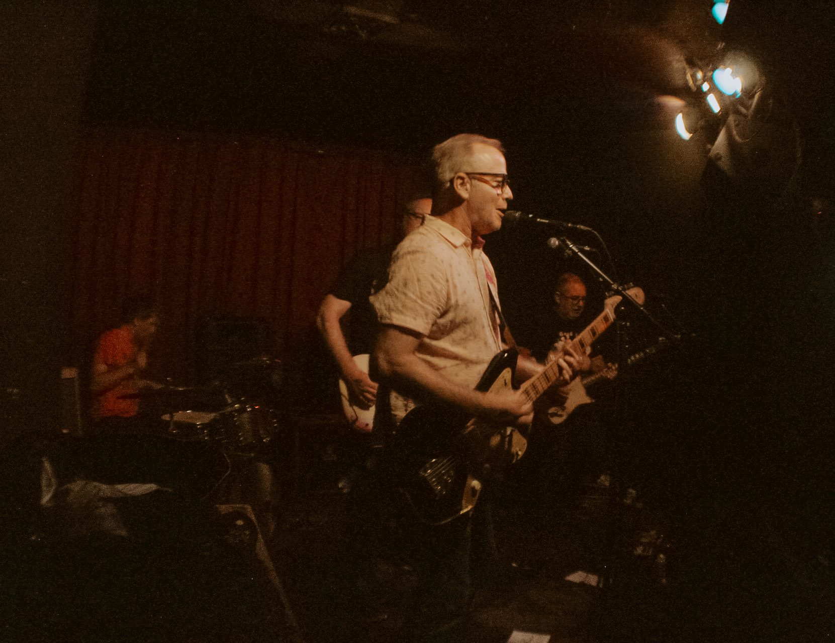 A live band performance featuring a guitarist singing into a microphone, with a drummer in the background and red curtains behind them.