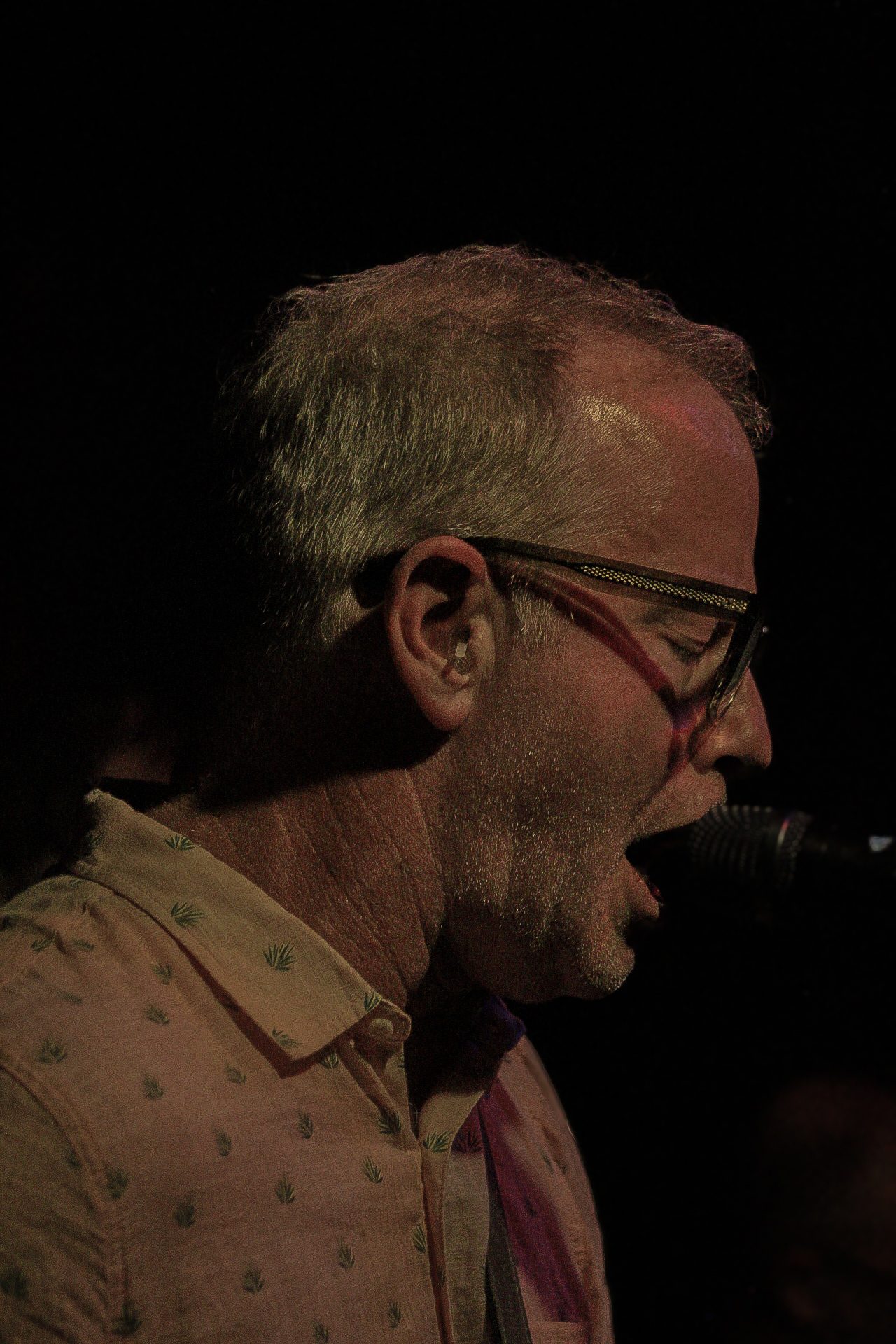 A close-up of a man singing into a microphone, wearing glasses and a patterned shirt, illuminated by stage lighting.