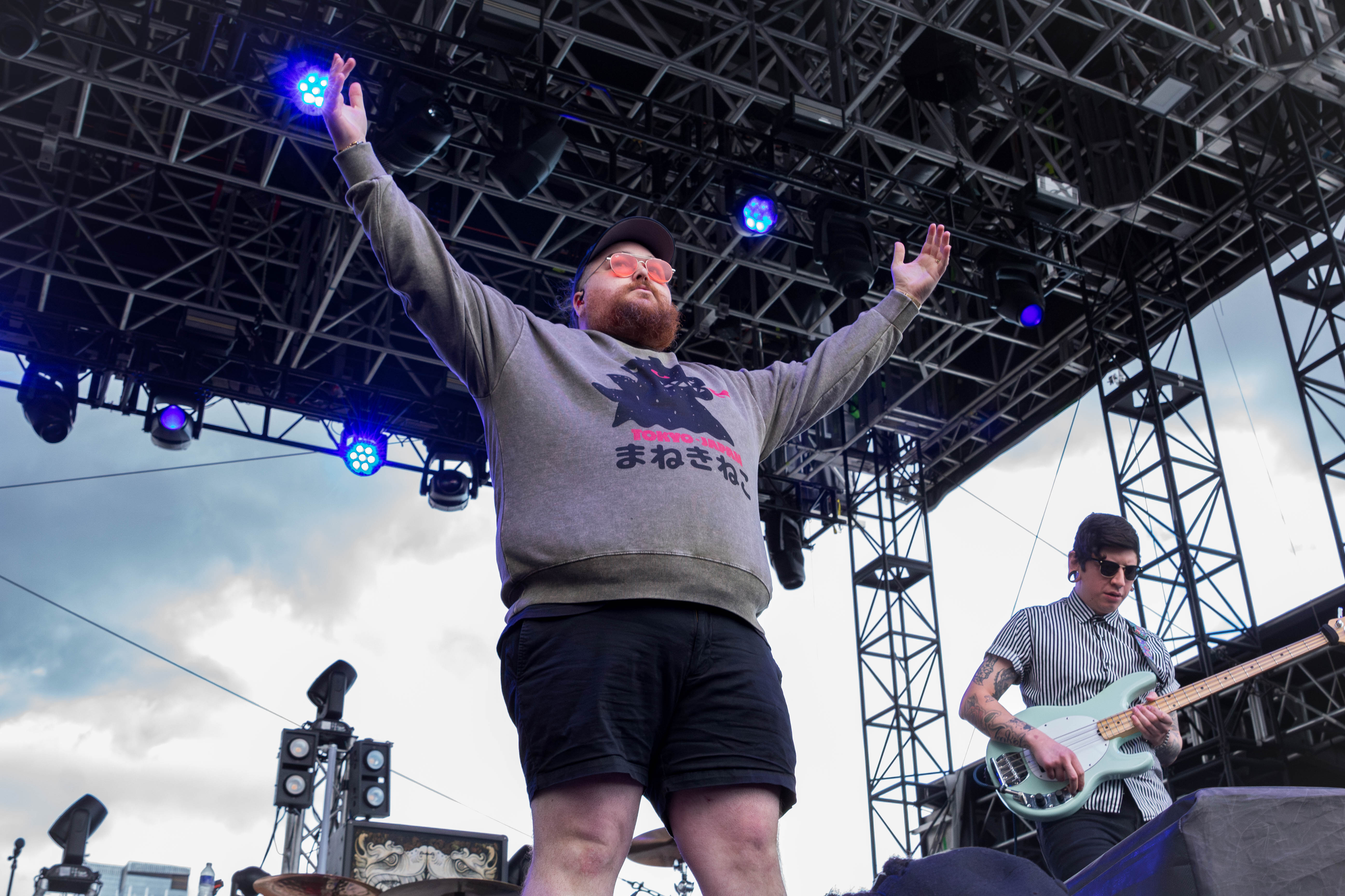 A performer with arms raised, wearing a gray sweater and sunglasses, is on stage during a live music event, while a musician plays bass in the background.