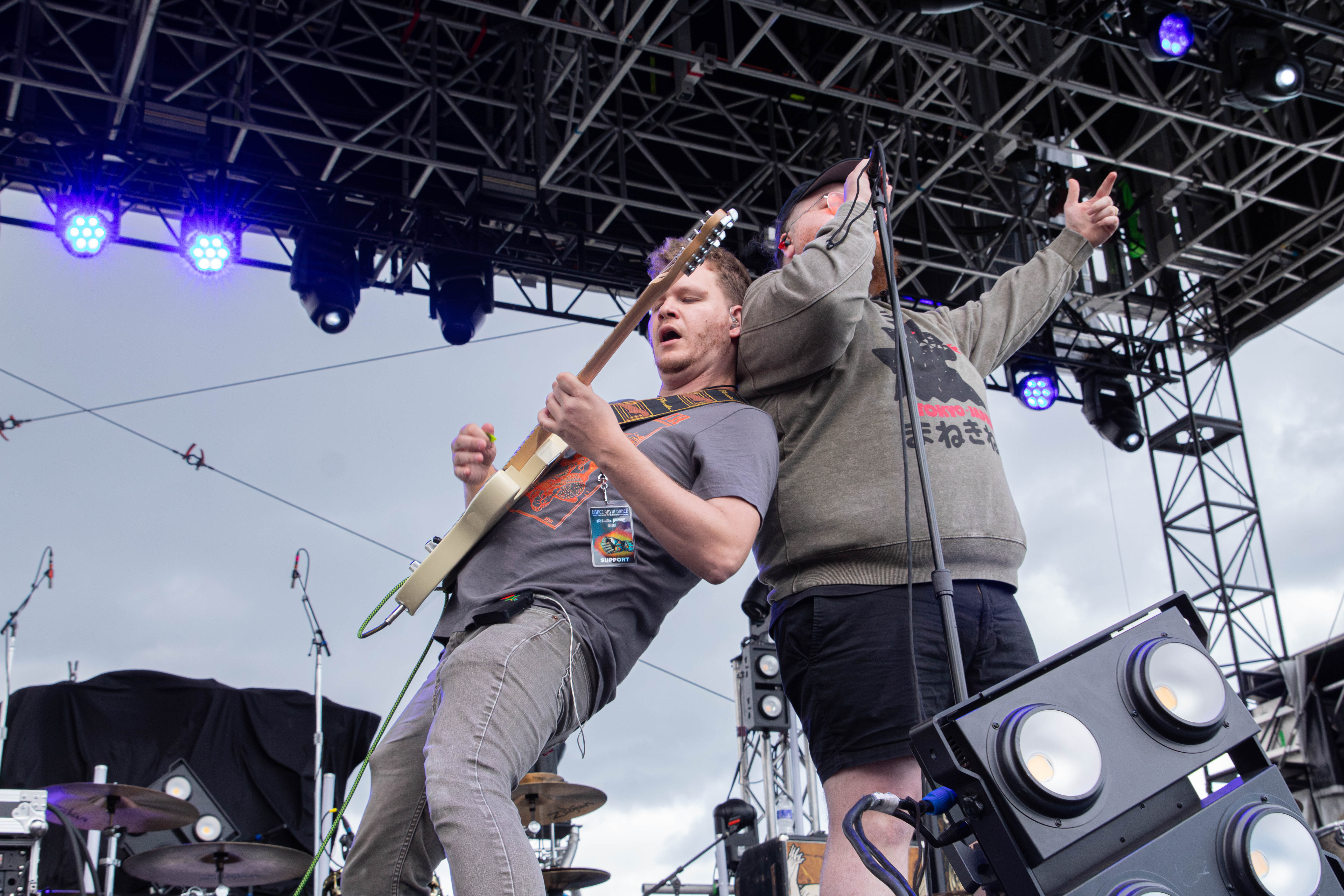 Two musicians performing on stage, one playing an electric guitar and the other singing into a microphone, with a concert lighting setup in the background.