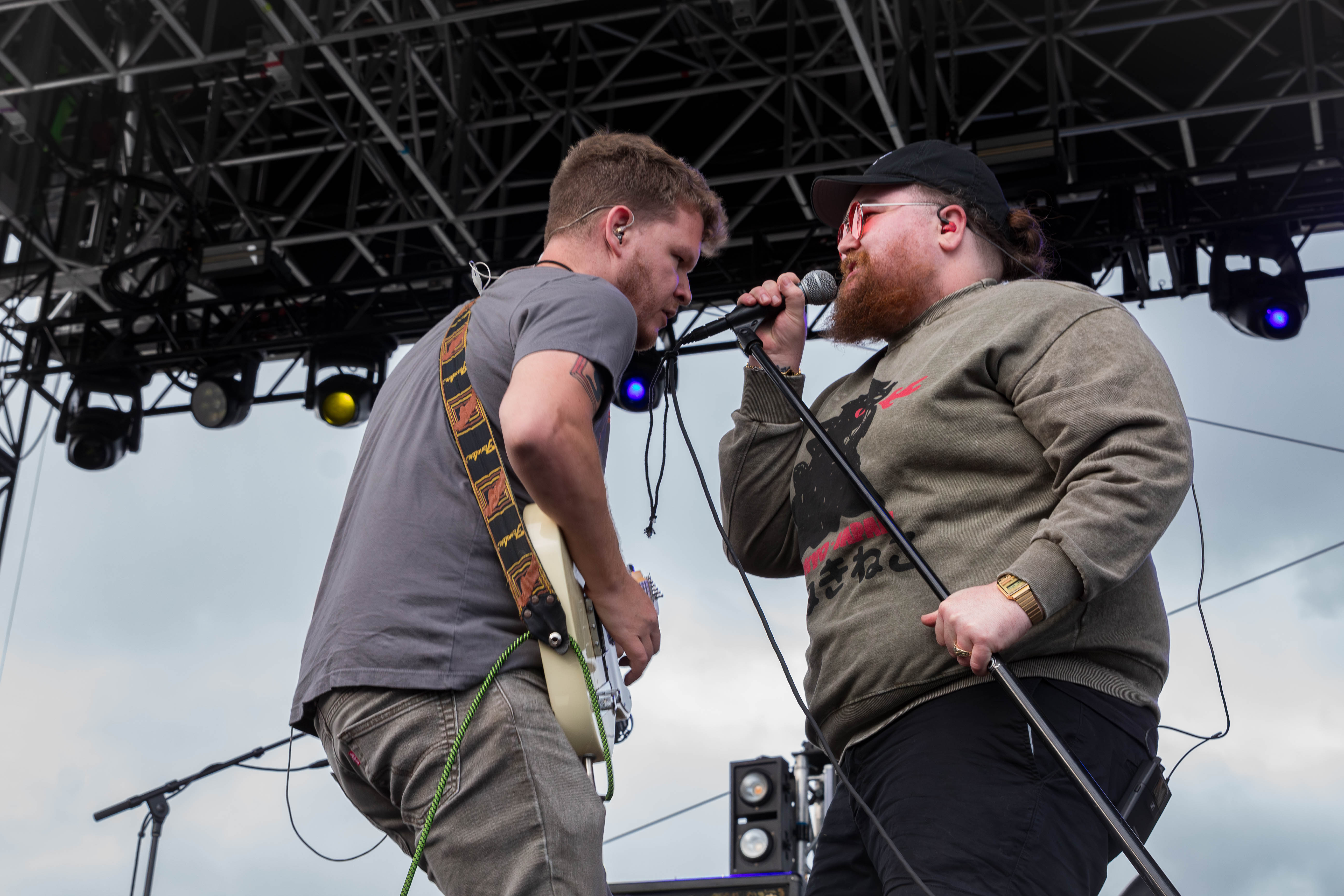 Two musicians performing on stage, one playing guitar and the other singing into a microphone, with a concert setting and lighting in the background.