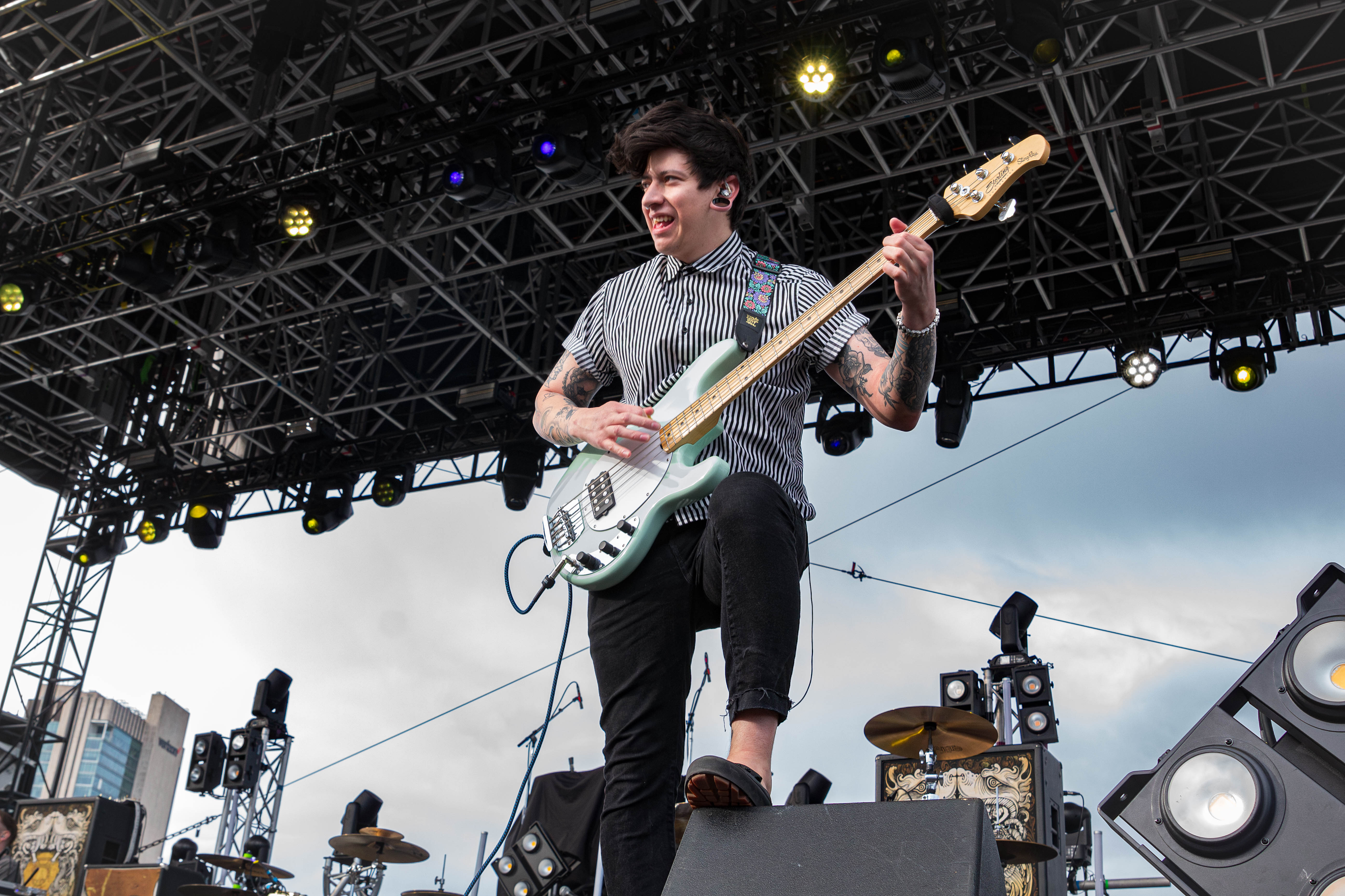 A musician playing a bass guitar on stage during a concert. He has a playful expression and is wearing a striped shirt. The background features stage lights and equipment.