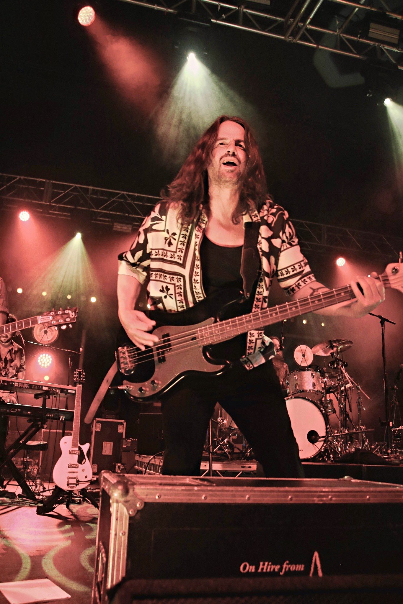 A musician passionately performing on stage while playing a bass guitar, wearing a patterned shirt, with dramatic lighting in the background.