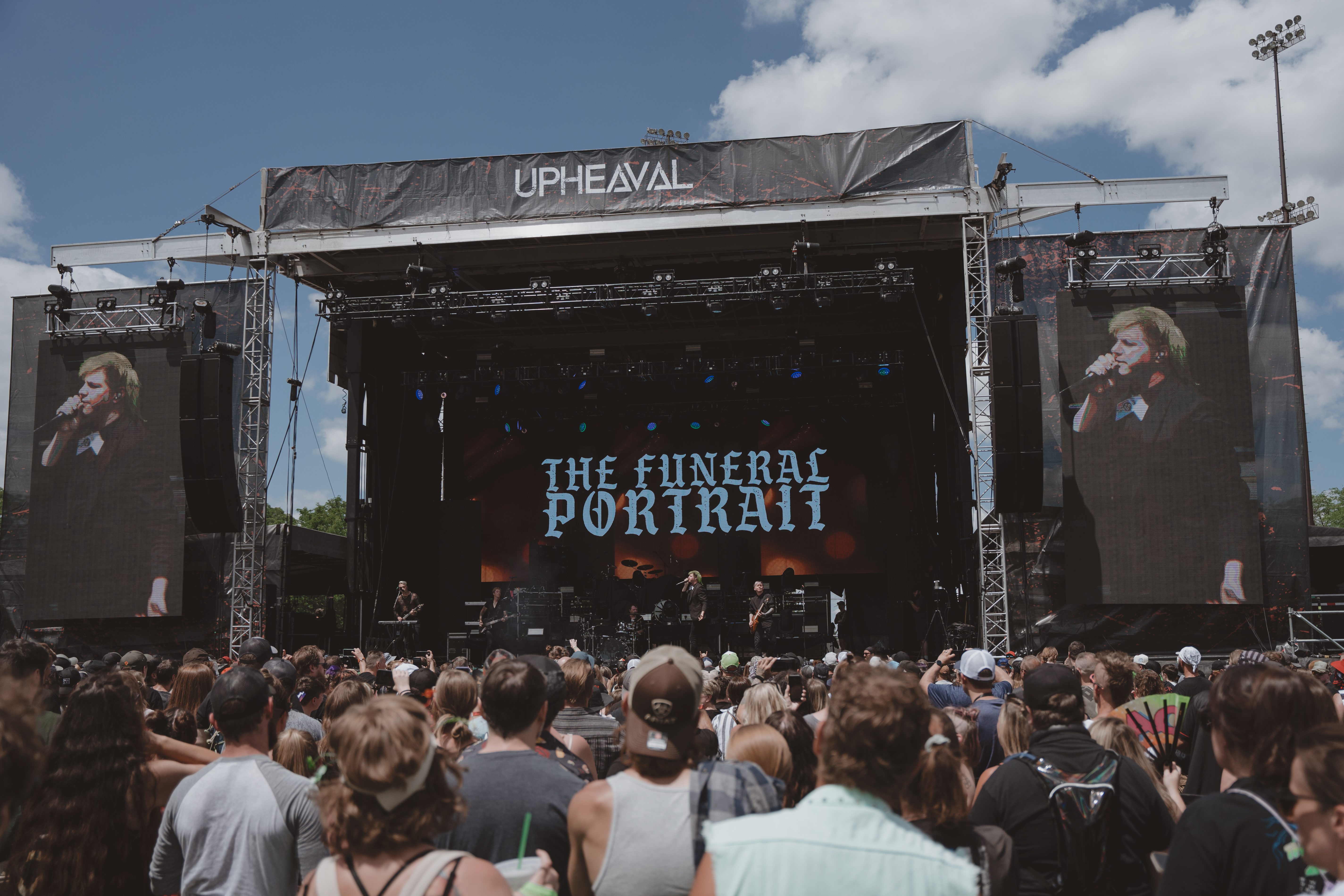 A live performance by The Funeral Portrait at the Upheaval Festival, with a large crowd in front of the stage under a clear blue sky.