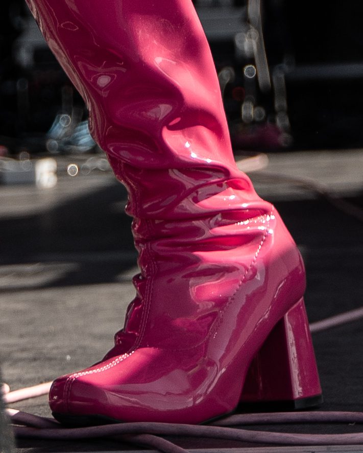 Close-up of a shiny pink high-heeled boot on stage, showcasing its glossy finish and dramatic silhouette.