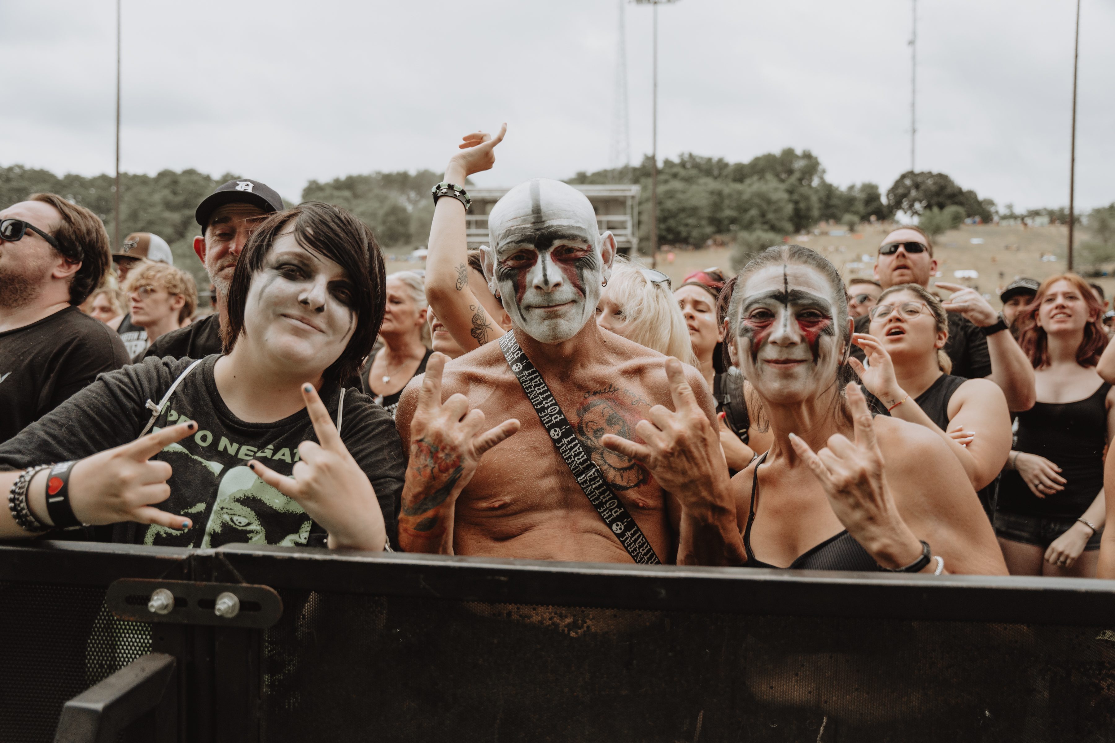 A lively festival crowd with attendees displaying face paint, including a person with black and white face paint and two individuals making rock hand gestures.