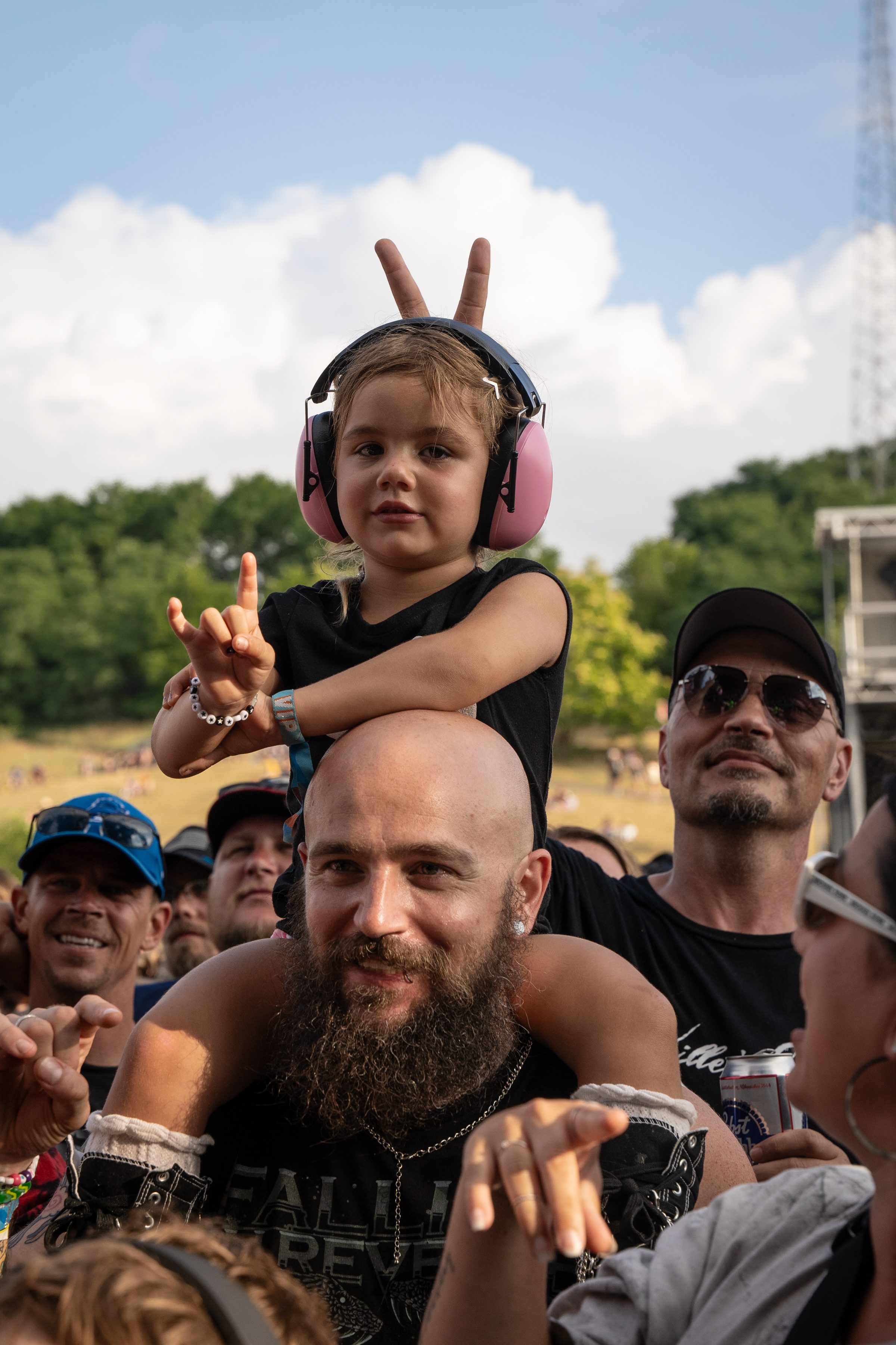 A young child with hearing protection headphones sits excitedly on a man's shoulders at a music festival, making rock hand gestures, surrounded by cheering fans.