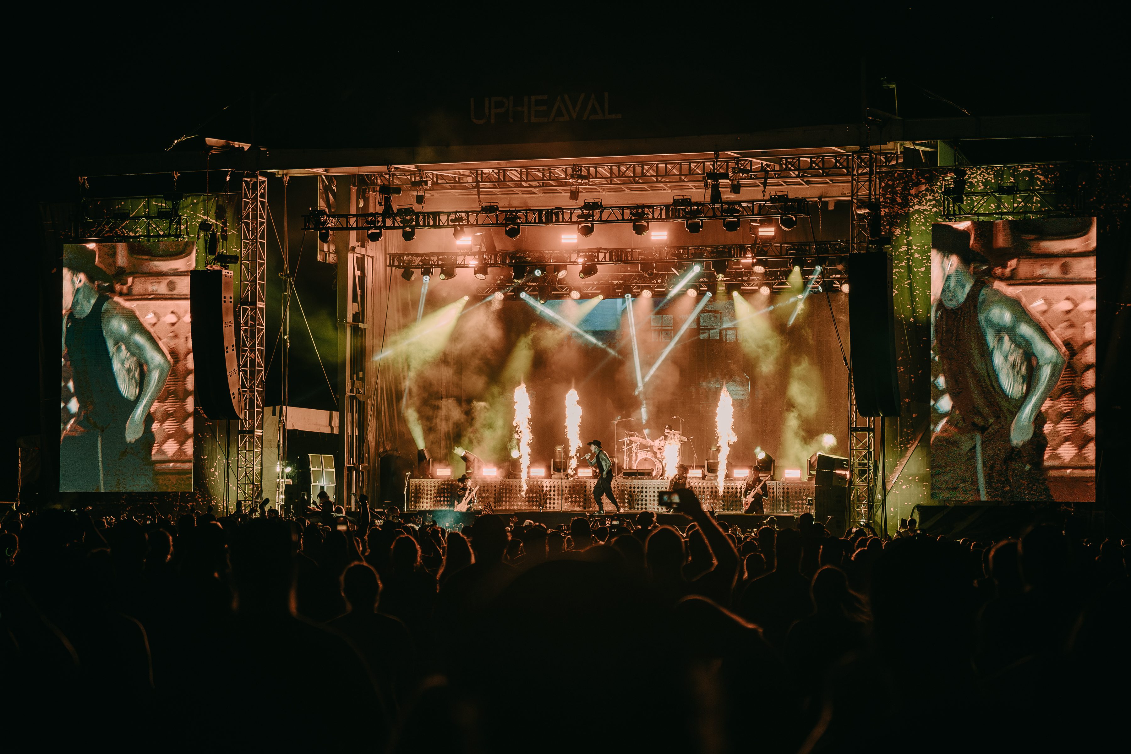 A live performance at the Upheaval Festival featuring a band on stage with vibrant lighting and pyrotechnics, surrounded by a large crowd enjoying the music.