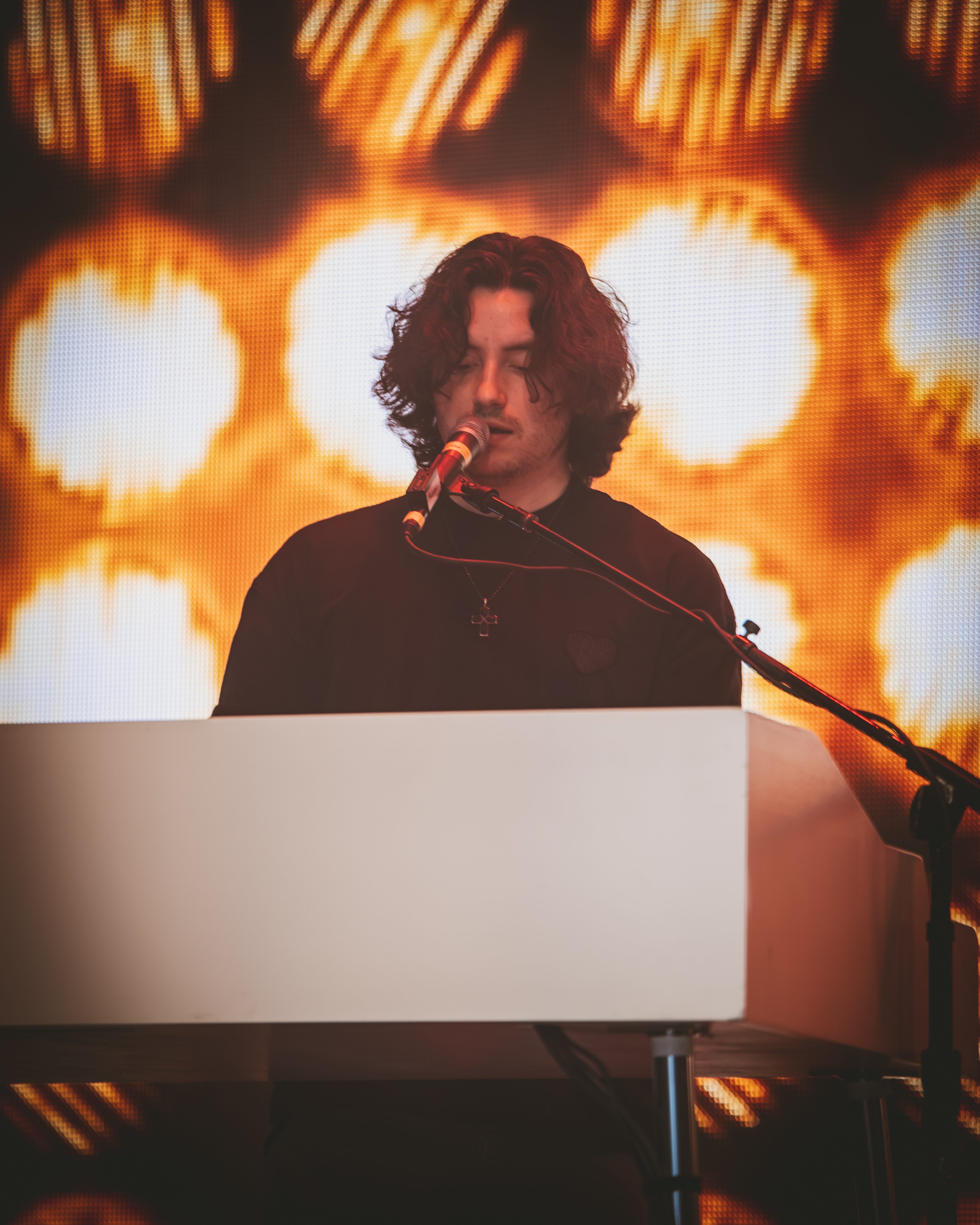 A young musician performing on stage at a concert, standing behind a keyboard with a microphone, illuminated by bright, warm lights in the background.