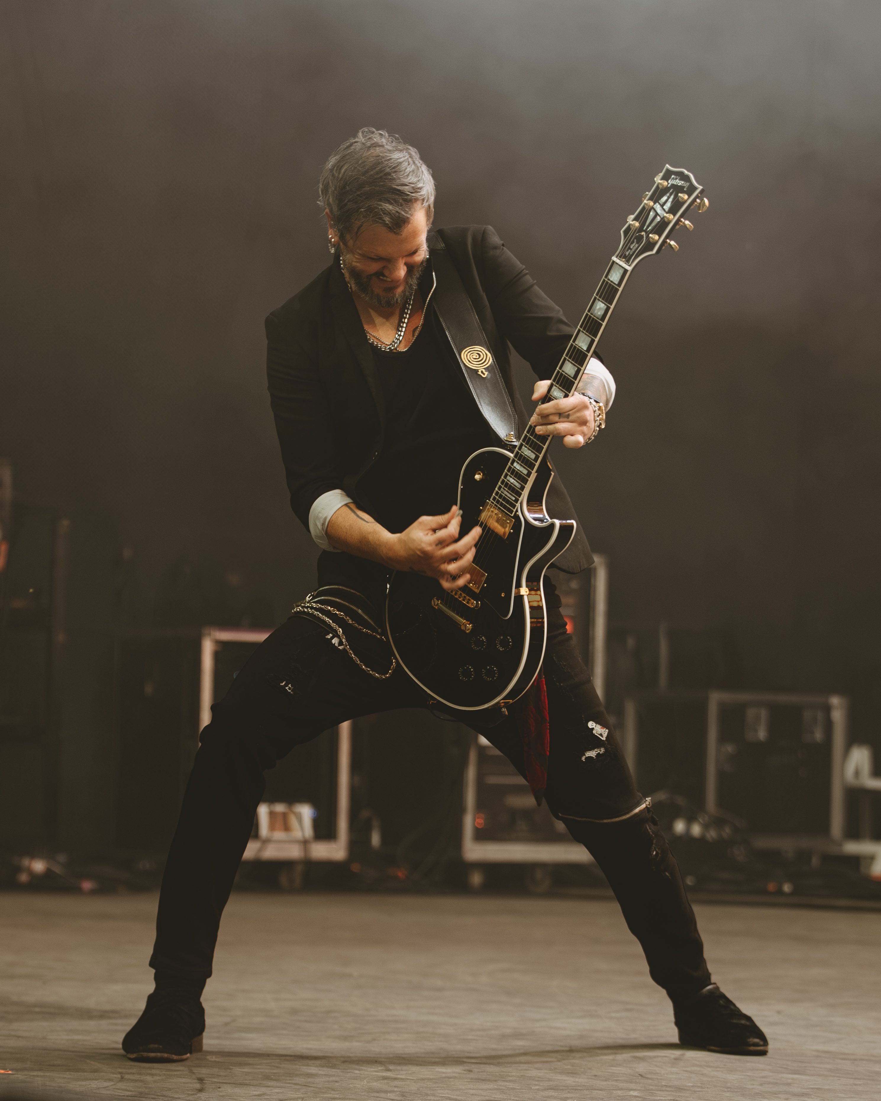 A guitarist passionately performing on stage, playing a black electric guitar with a dramatic stance as smoke fills the background.