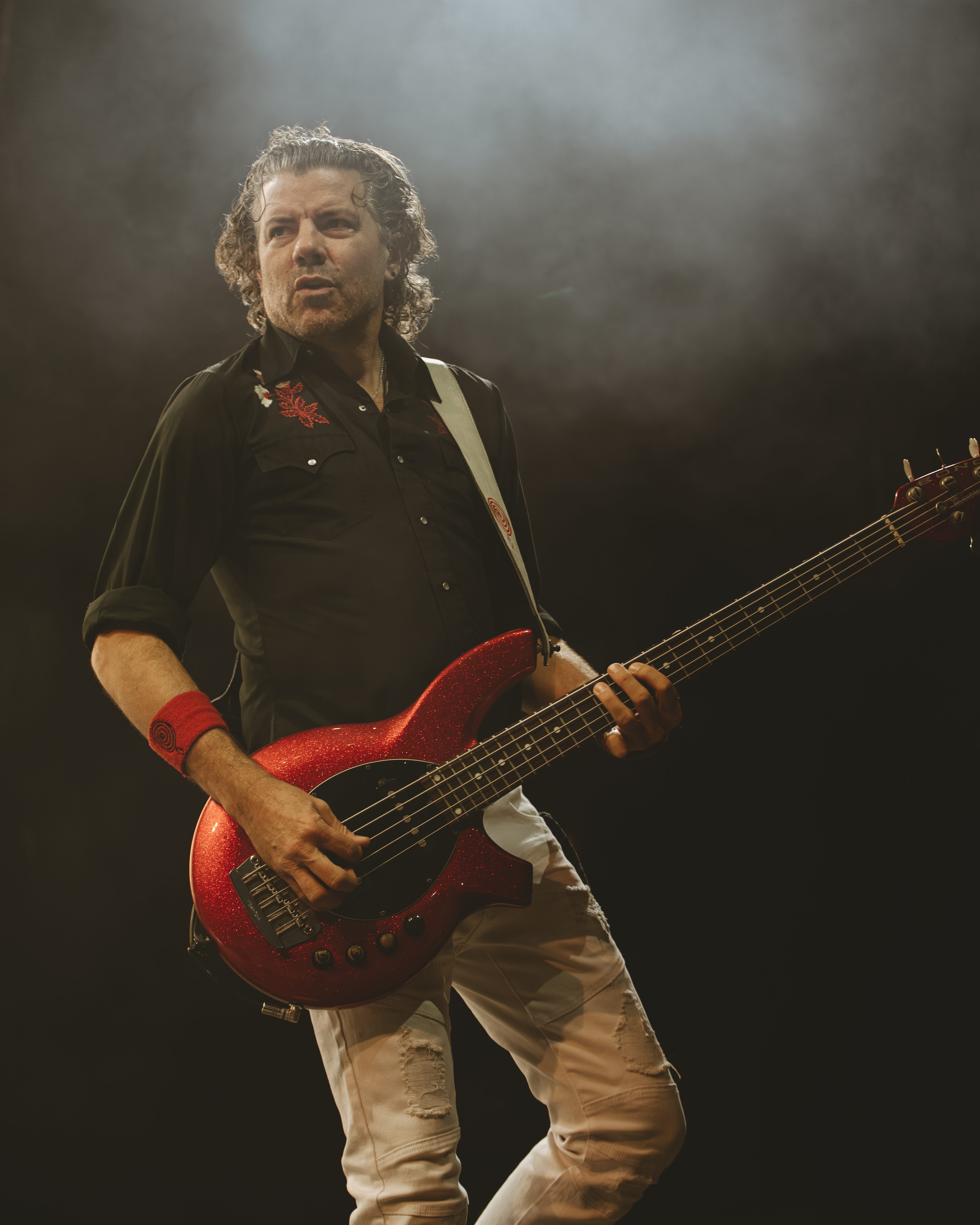 A musician performing on stage with a sparkling red bass guitar, wearing a black shirt and white pants, surrounded by a smoky atmosphere.