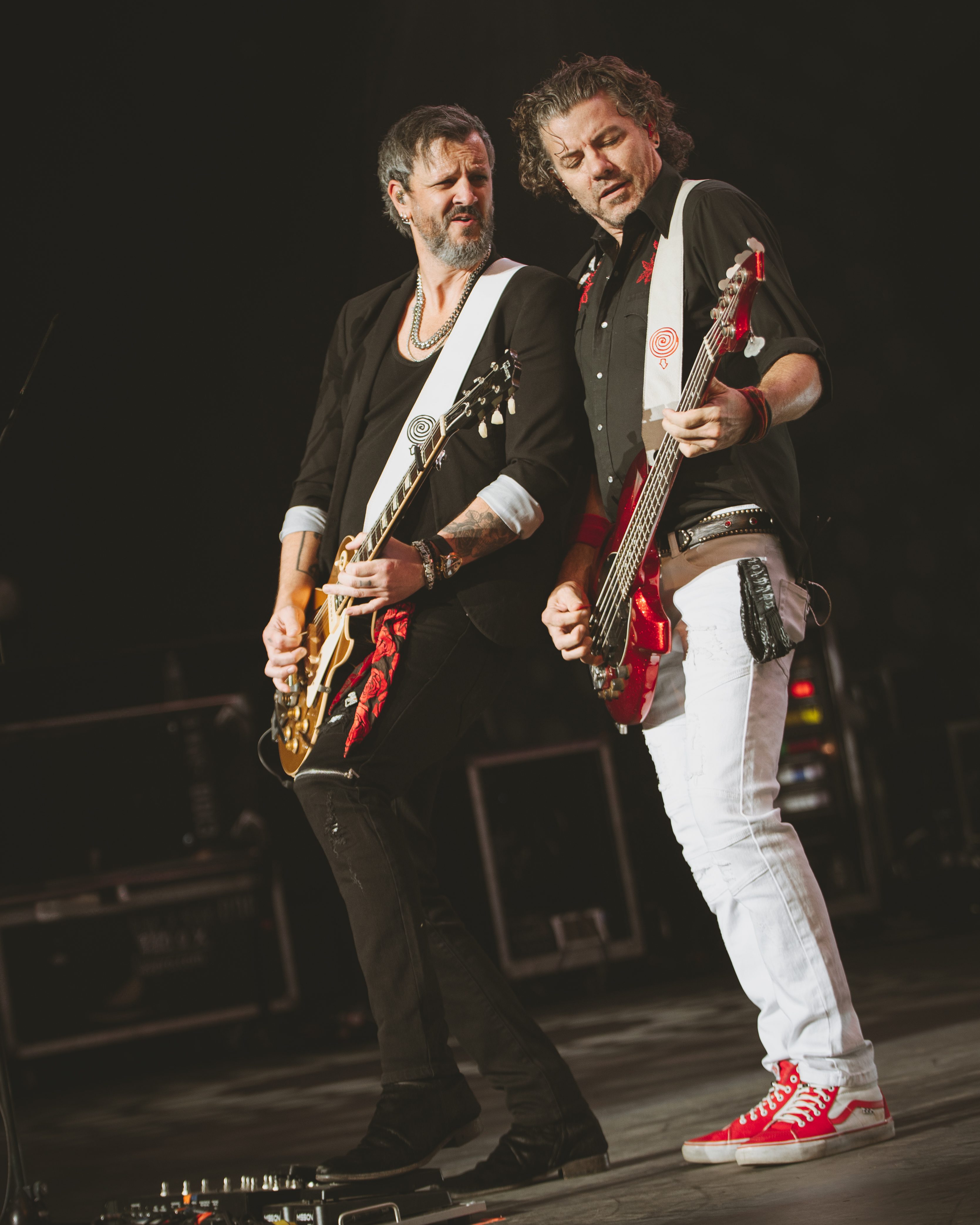 Two rock musicians performing on stage, one playing a guitar and the other a bass guitar, both wearing stylish outfits.