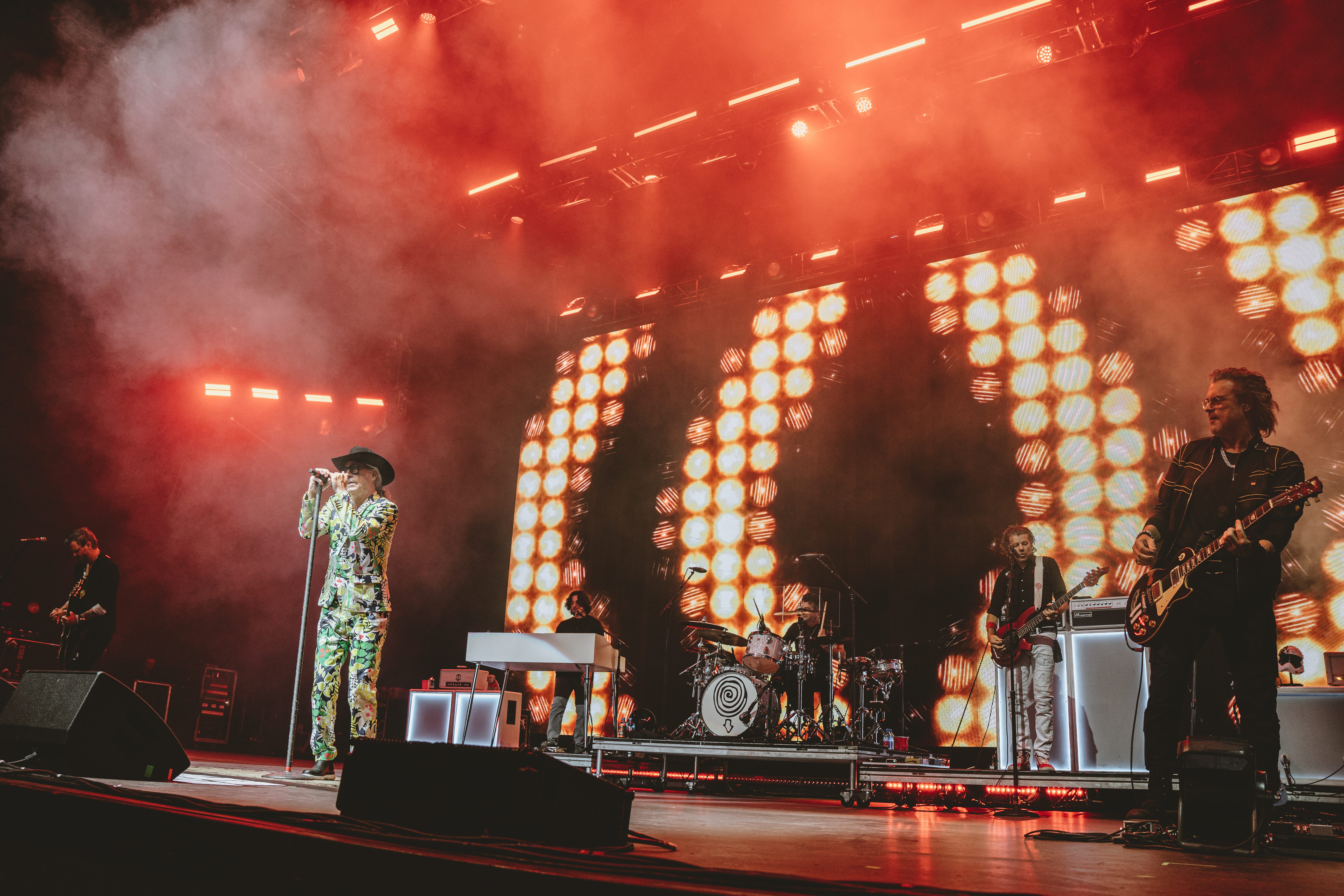 A band performing live on stage with vibrant lighting and smoke effects, featuring a lead vocalist in a colorful patterned suit and other band members playing instruments.