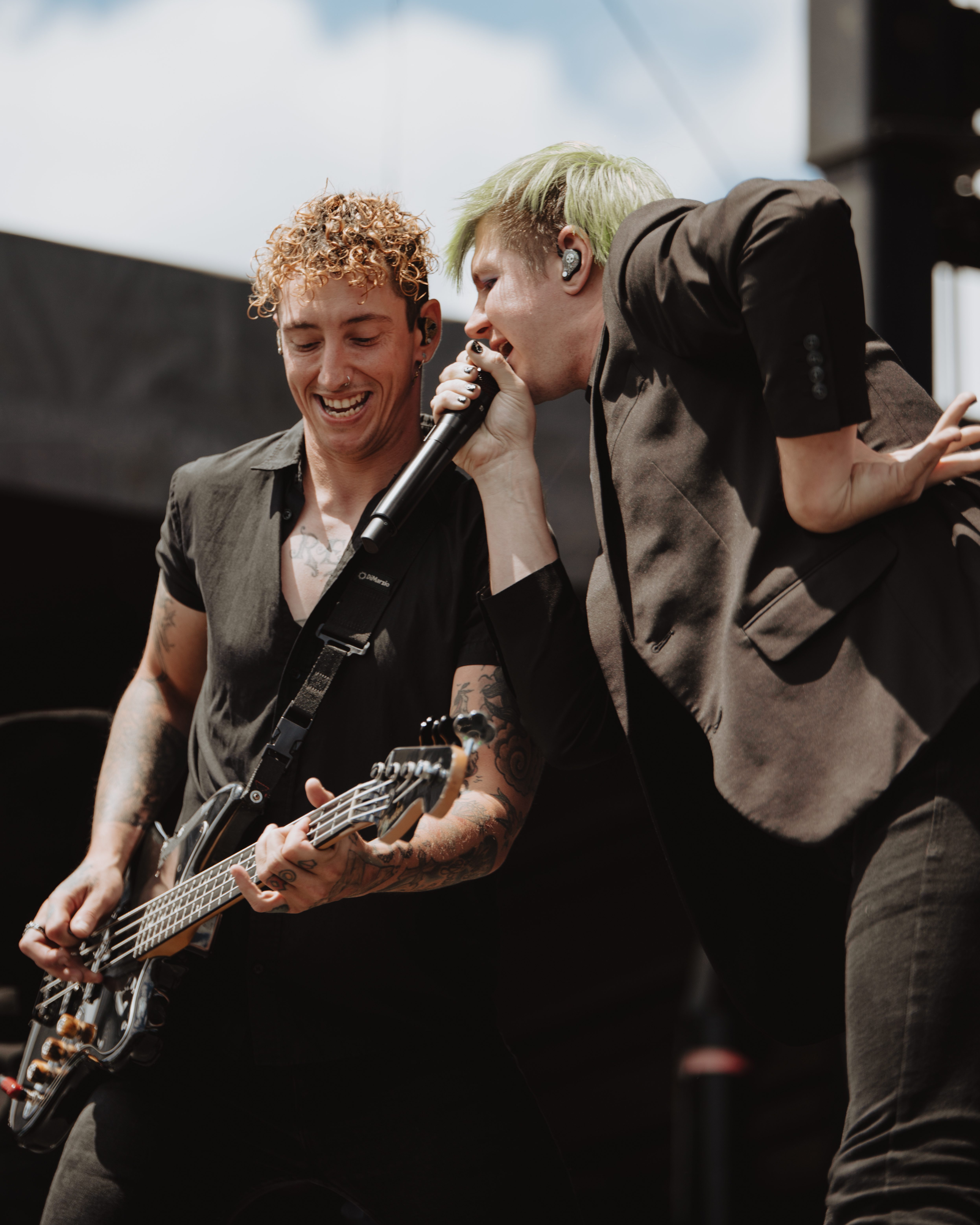 Two male musicians performing on stage, one playing the guitar and the other singing into a microphone, both smiling and engaged in their performance under a clear blue sky.