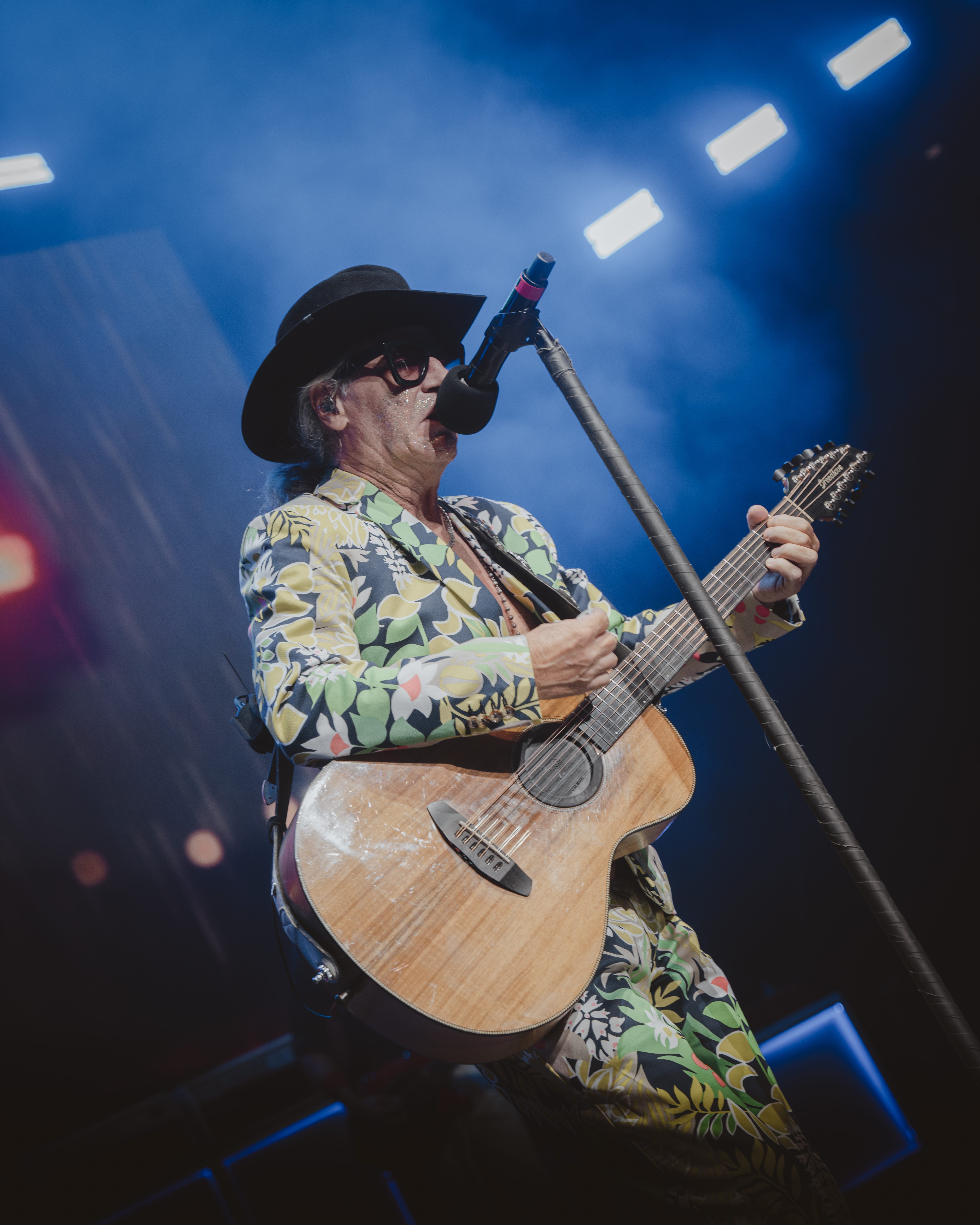 A musician wearing a colorful floral suit and a cowboy hat performs on stage while playing an acoustic guitar and singing into a microphone, with a smoky blue background.