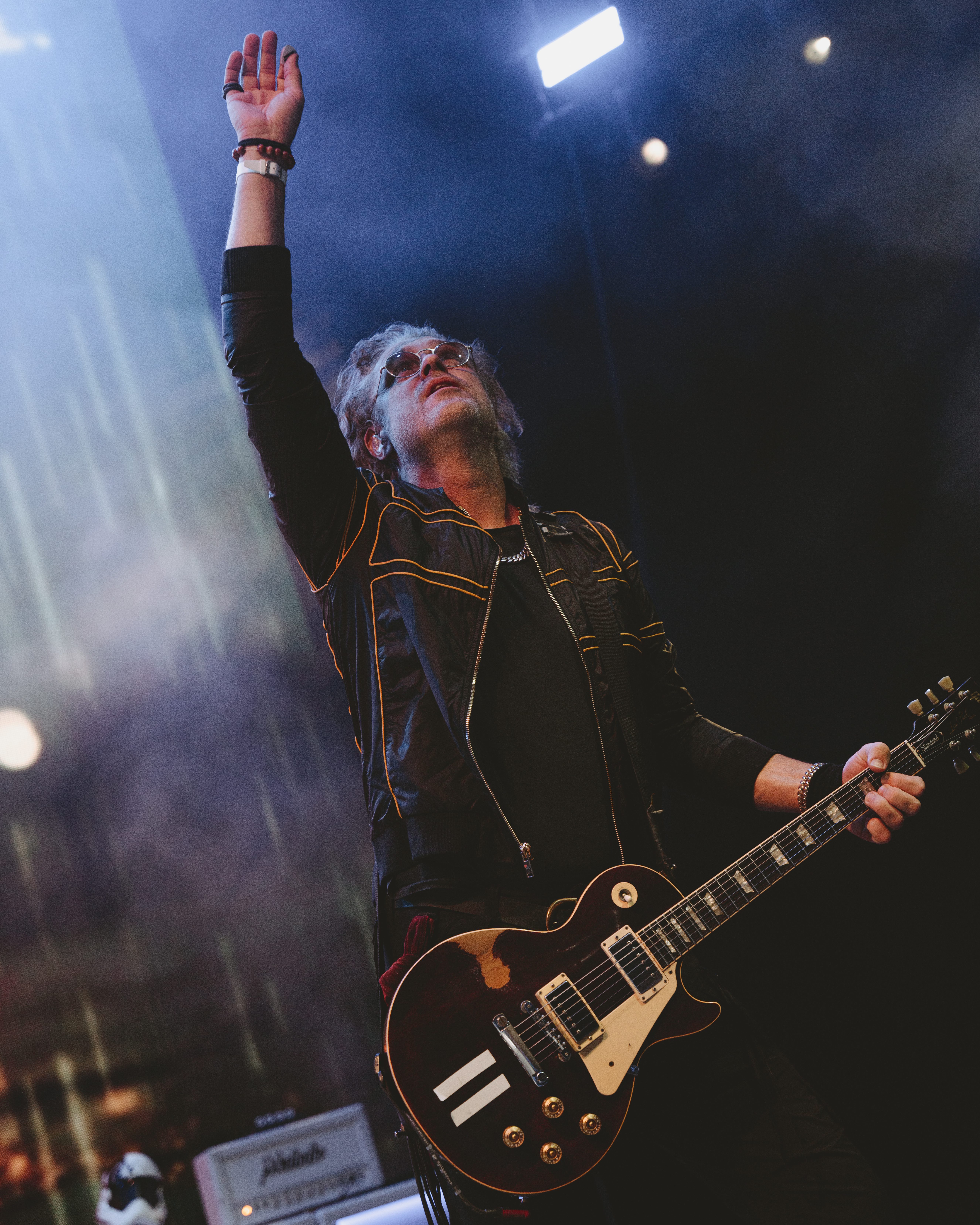 A guitarist performing on stage with his arm raised, wearing a black jacket with yellow accents, set against a smoky background with stage lights.