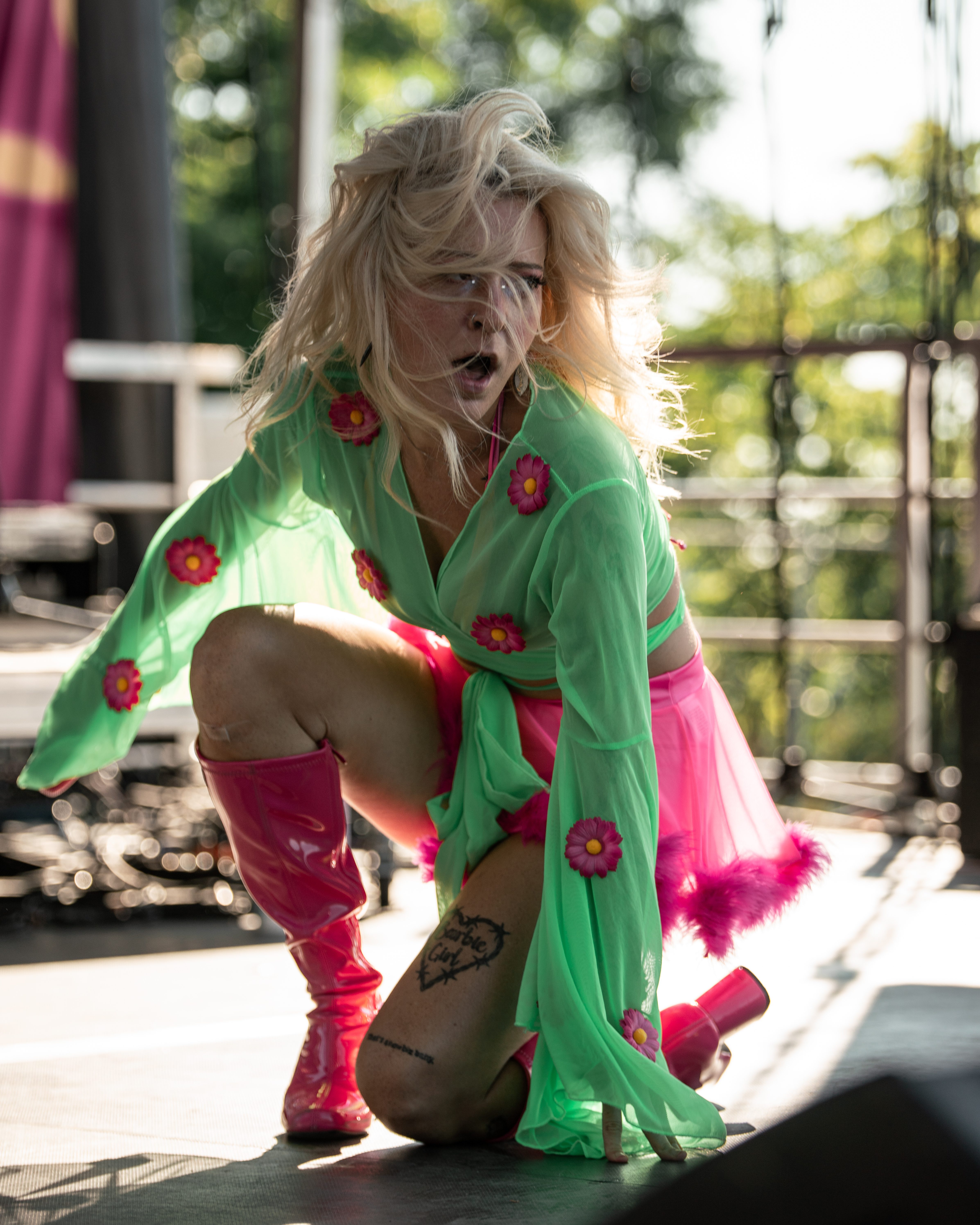 A performer in a vibrant green outfit adorned with pink flowers and a pink skirt crouches on stage, expressing dynamic energy during a live performance.