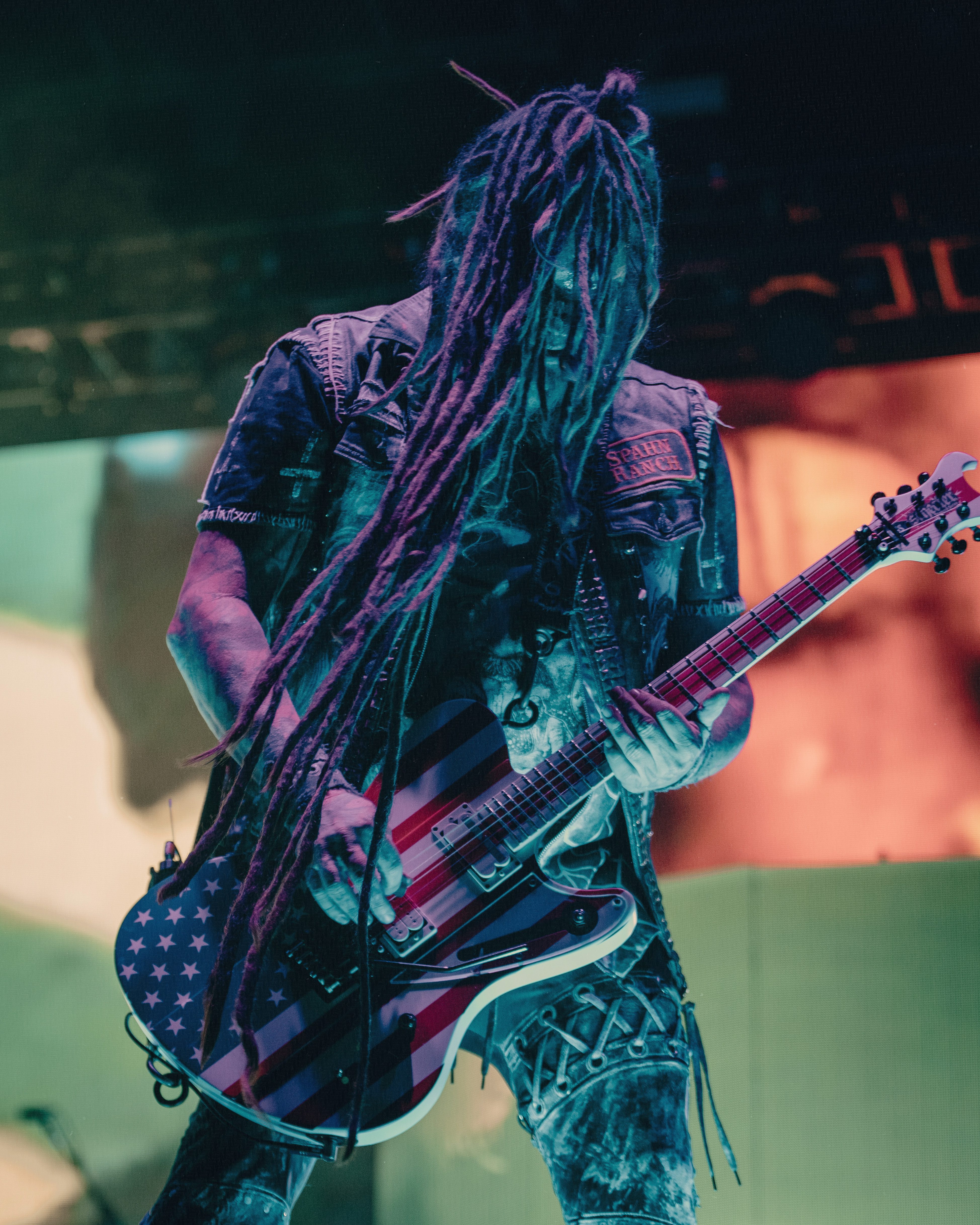 A musician with long dreadlocks performs on stage, holding an electric guitar adorned with an American flag design, against a colorful backdrop of stage lights.