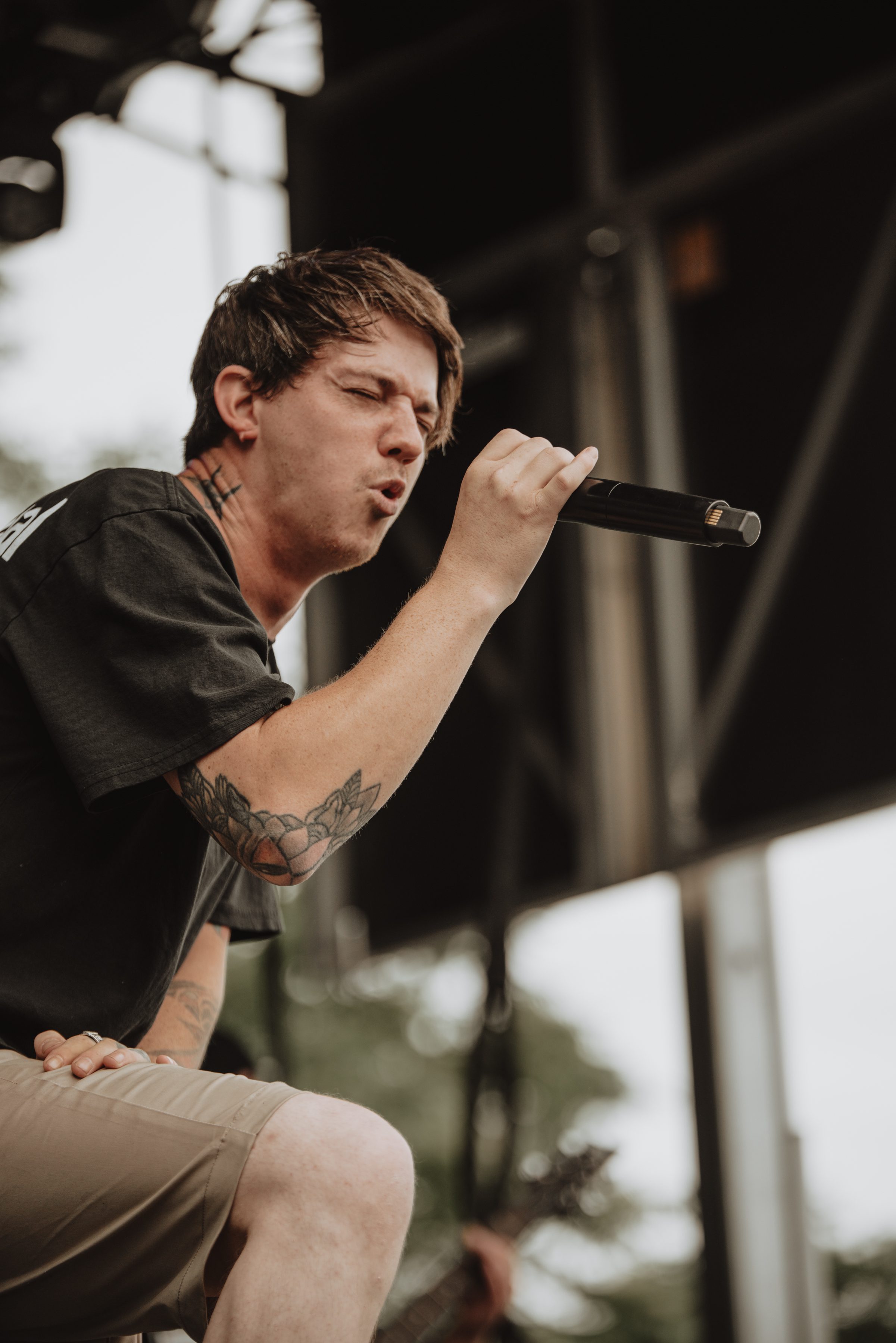 A male lead singer passionately performing on stage with a microphone, wearing a black t-shirt and shorts, showcasing visible tattoos on his arm.