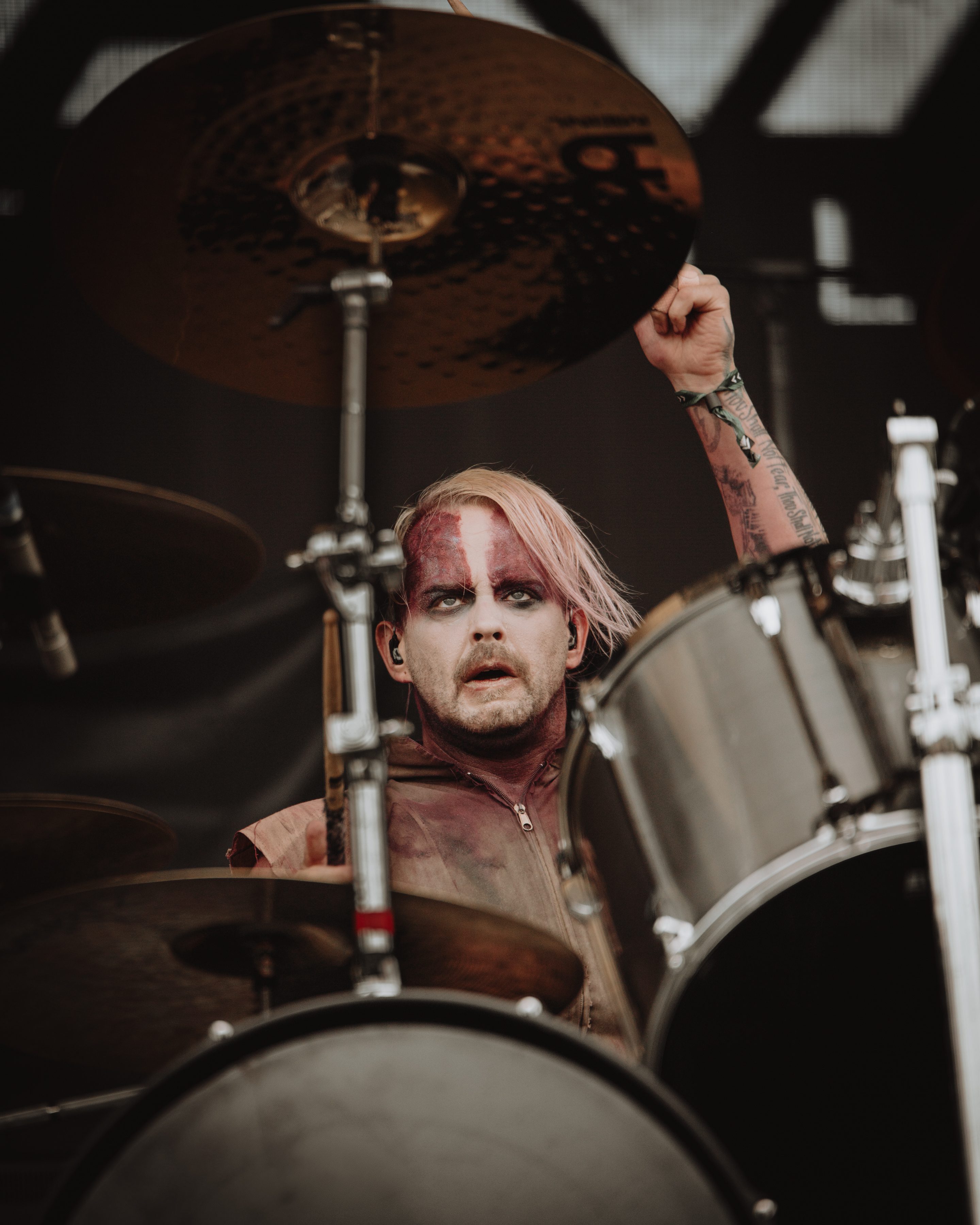 A drummer with pink hair and dramatic makeup passionately performing on stage, playing the drums with intensity.