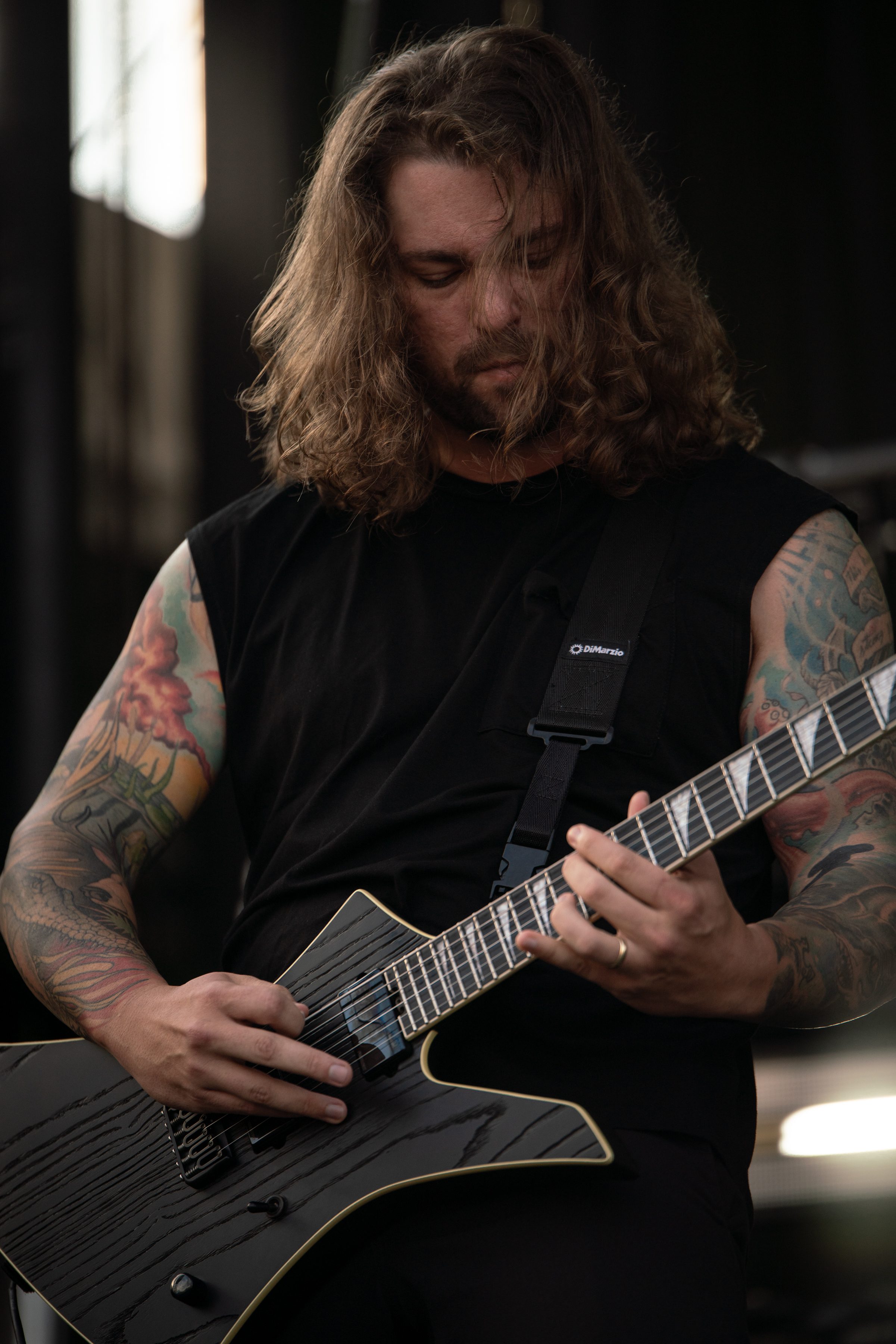A guitarist with long, wavy hair and tattooed arms passionately plays a black electric guitar on stage.