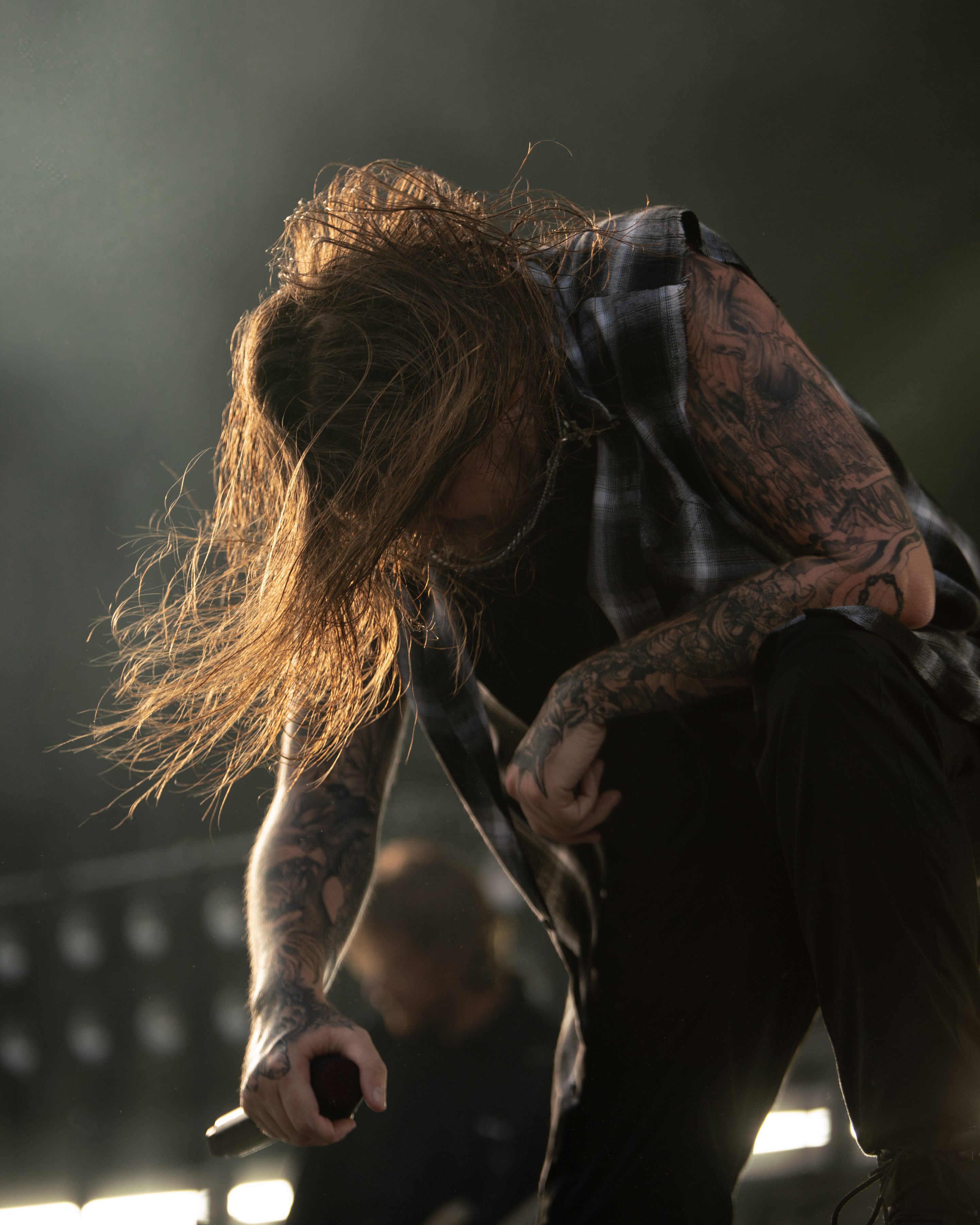 Musician with long hair and tattoos passionately performing on stage, holding a microphone while engaged in an energetic moment.