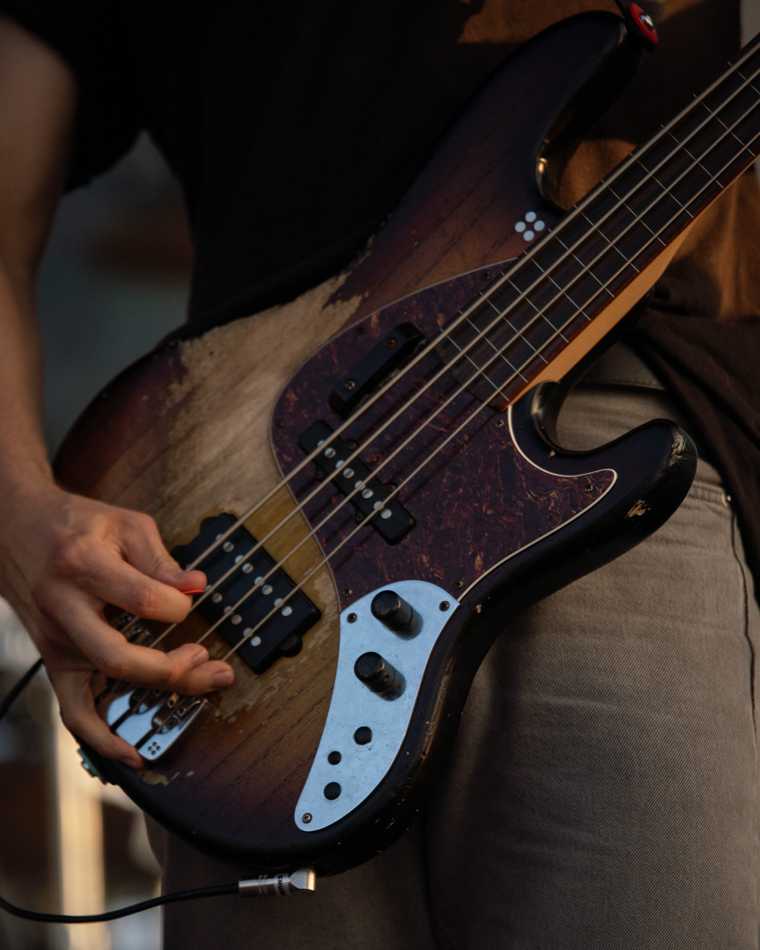 Close-up of a hand playing an electric bass guitar, showing the instrument's weathered body and control knobs, with a blurred background of a performance setting.