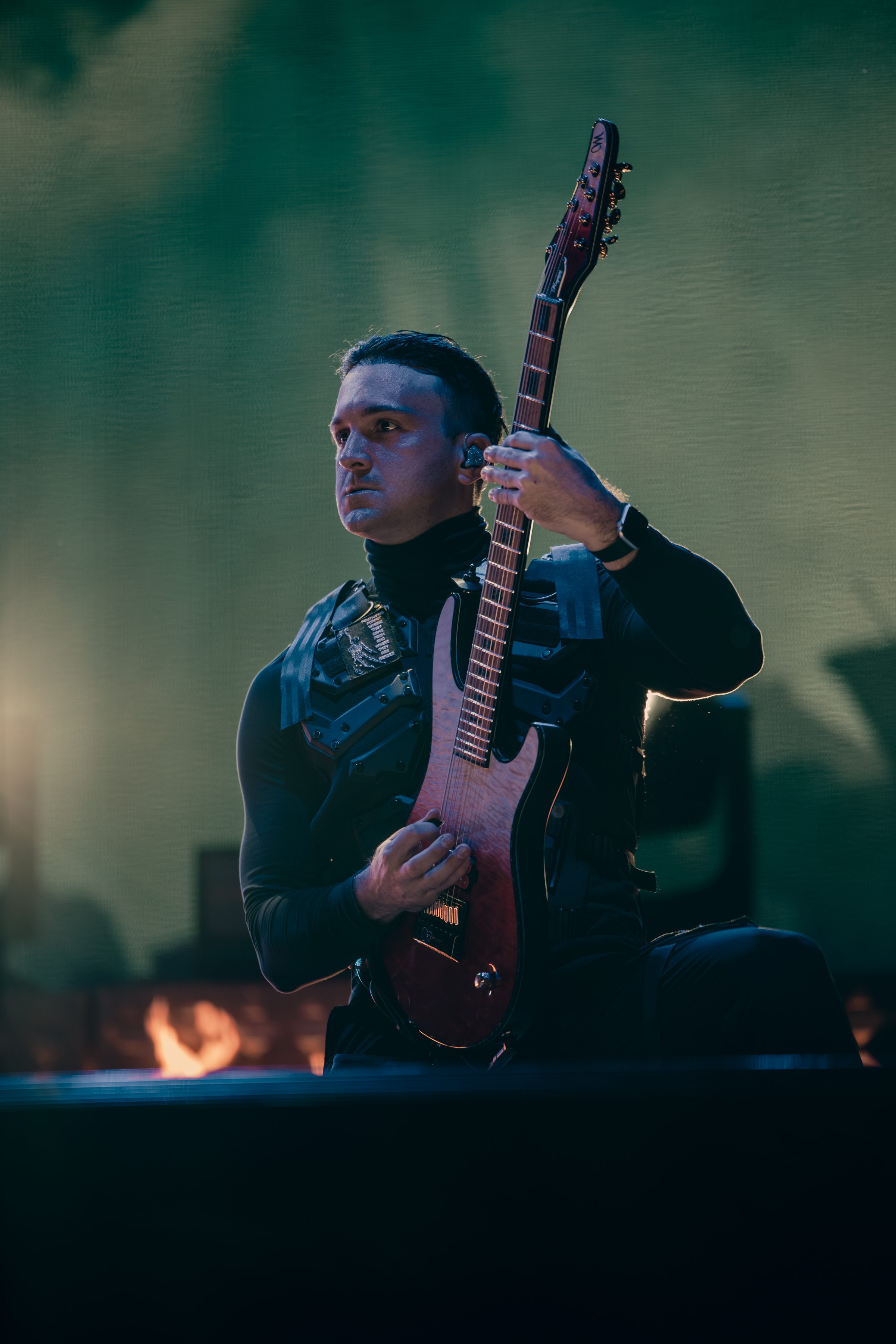 A musician in a black outfit plays an electric guitar on stage with a focused expression, illuminated by stage lights in the background.
