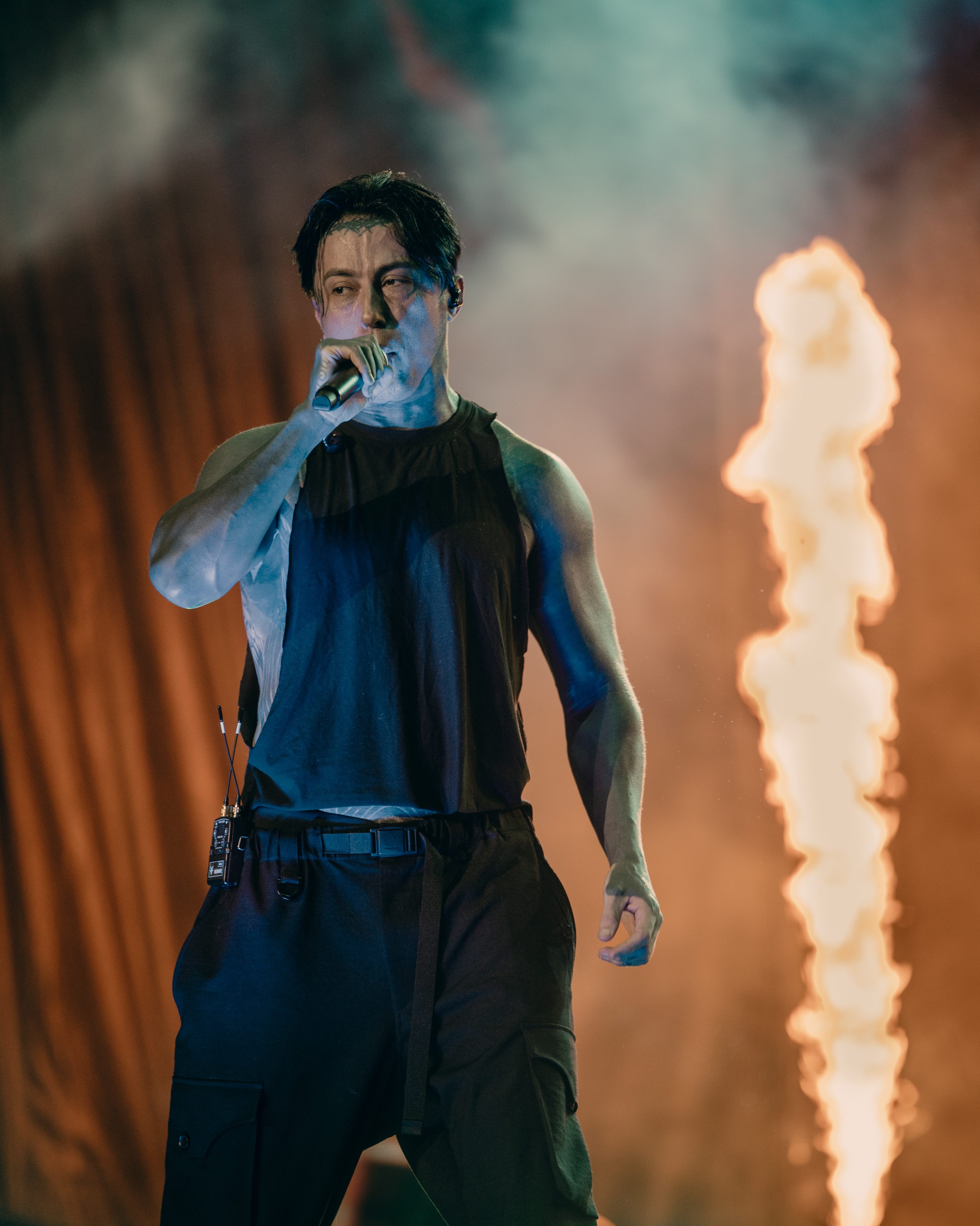 A male performer with dark hair and a sleeveless shirt passionately sings into a microphone on stage, with flames erupting behind him, creating a dramatic atmosphere.