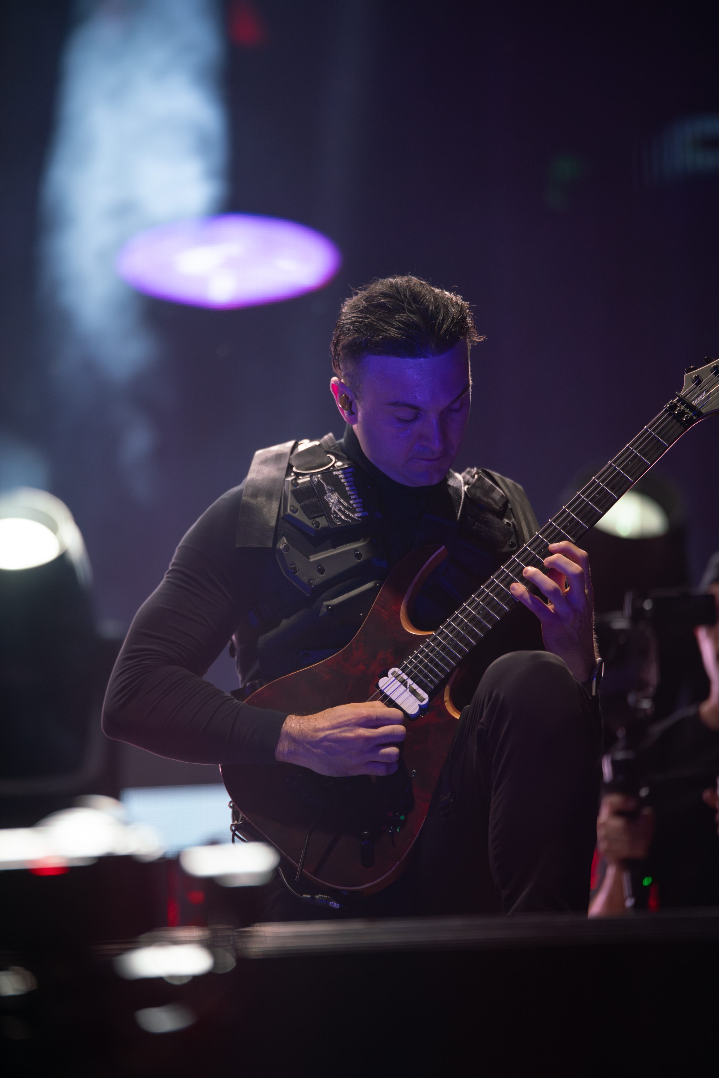 A musician performs on stage, focused on playing an electric guitar, wearing a black outfit and harness.