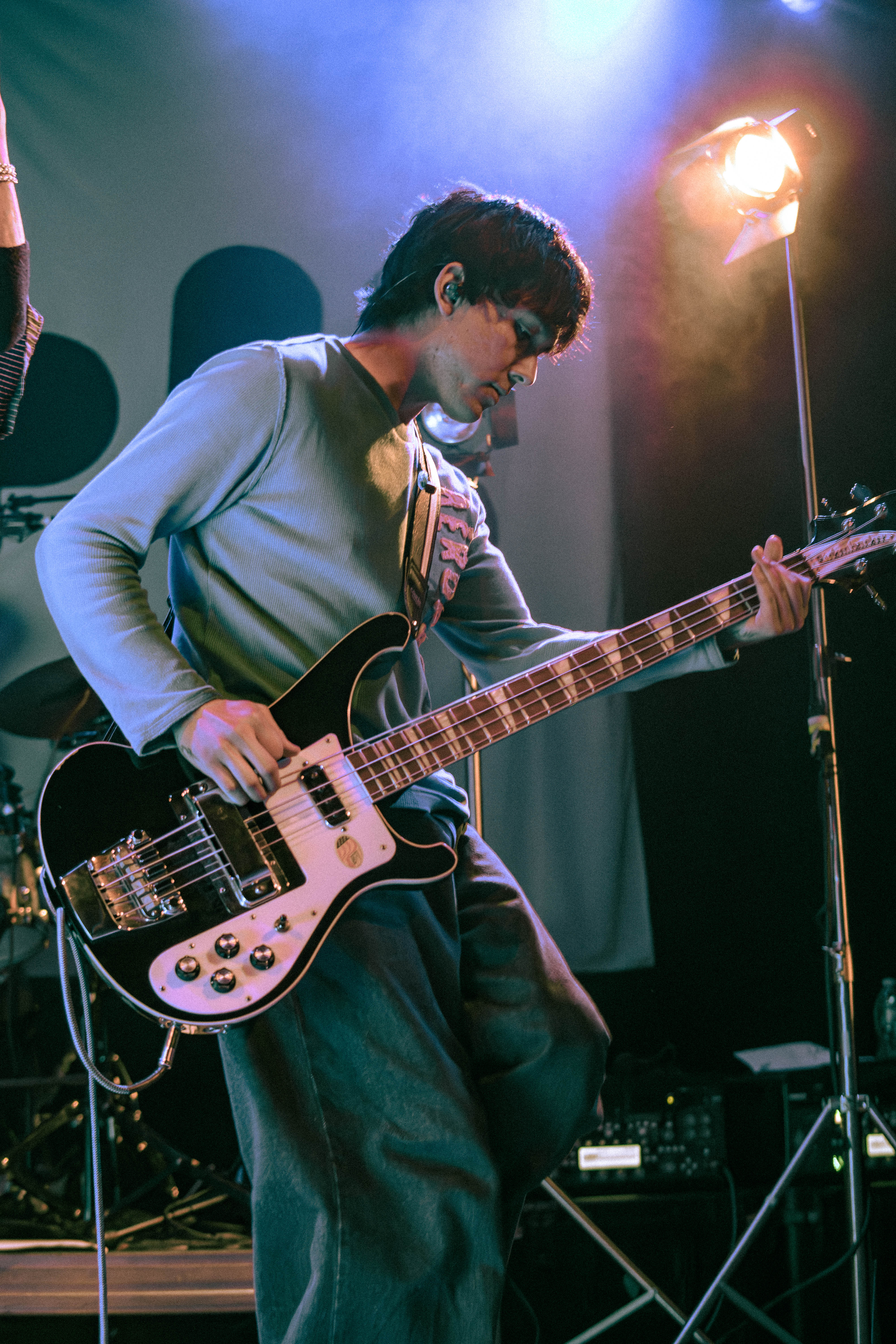 A musician playing an electric bass guitar on stage during a performance, with colorful stage lighting in the background.