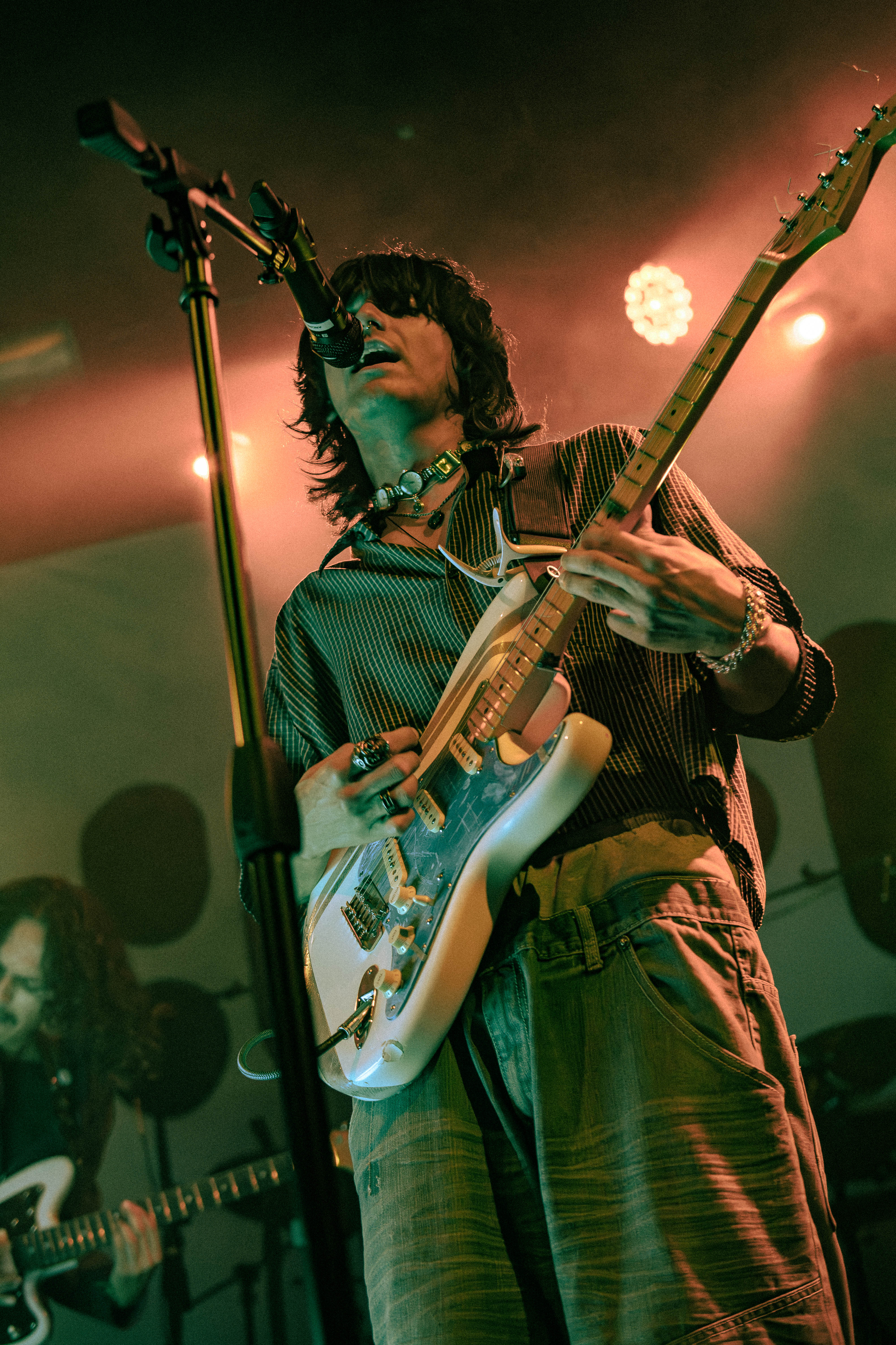 A musician performs on stage with a guitar, wearing a striped shirt and accessories, while another band member plays in the background under colorful lighting.