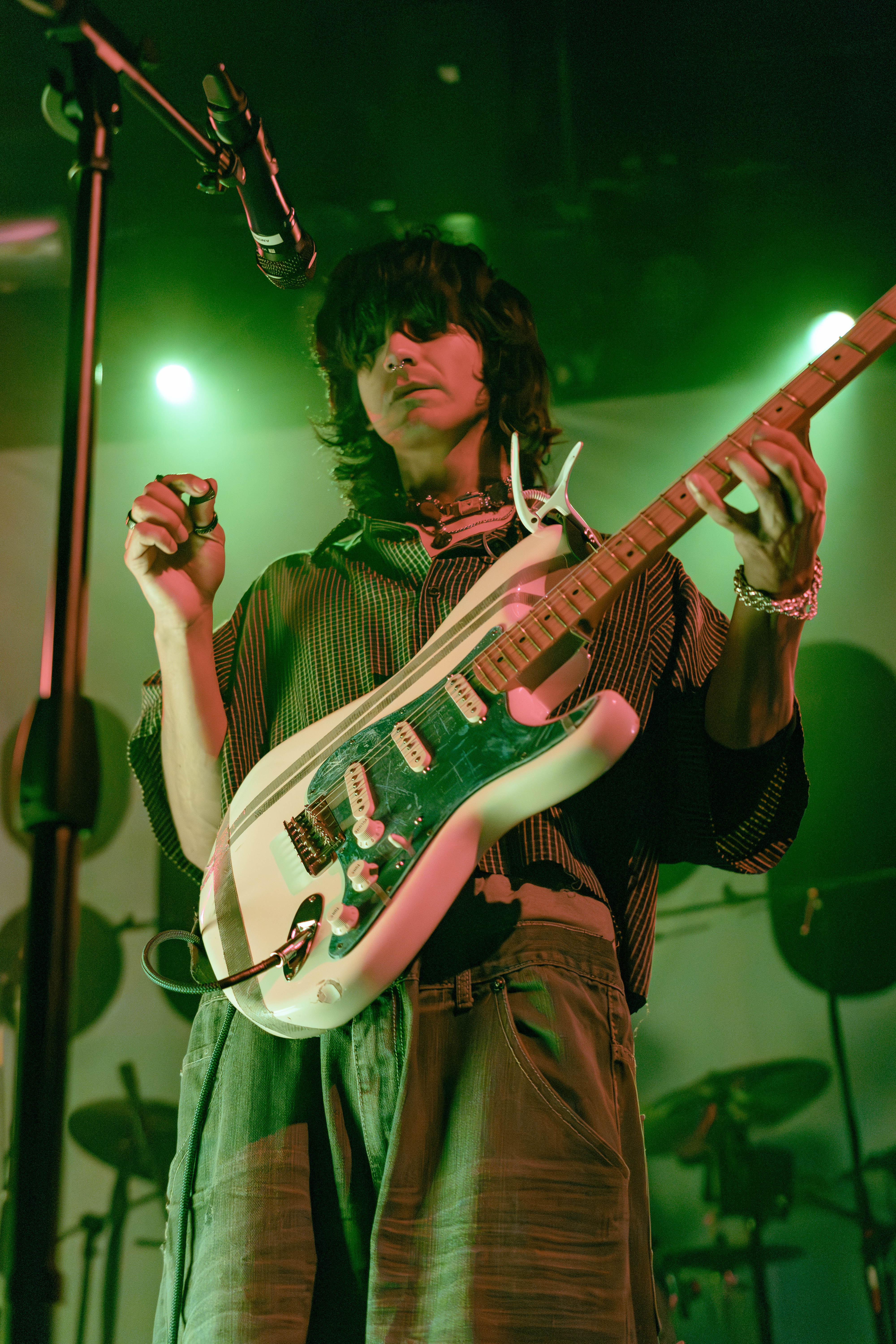 A musician performing on stage, holding an electric guitar and singing into a microphone, with green stage lighting illuminating the scene.