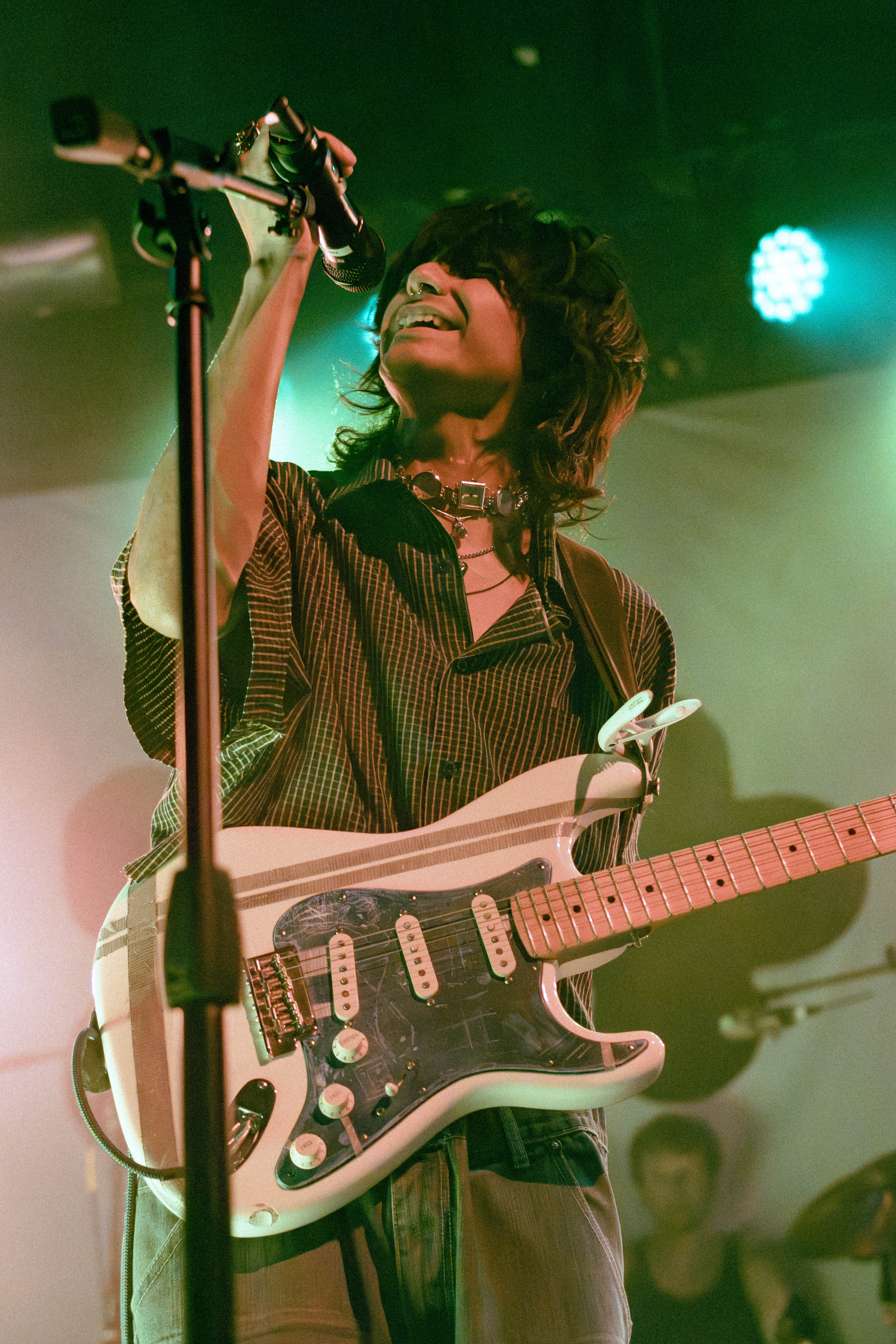 A musician performing on stage with a microphone and electric guitar, surrounded by soft green lighting.