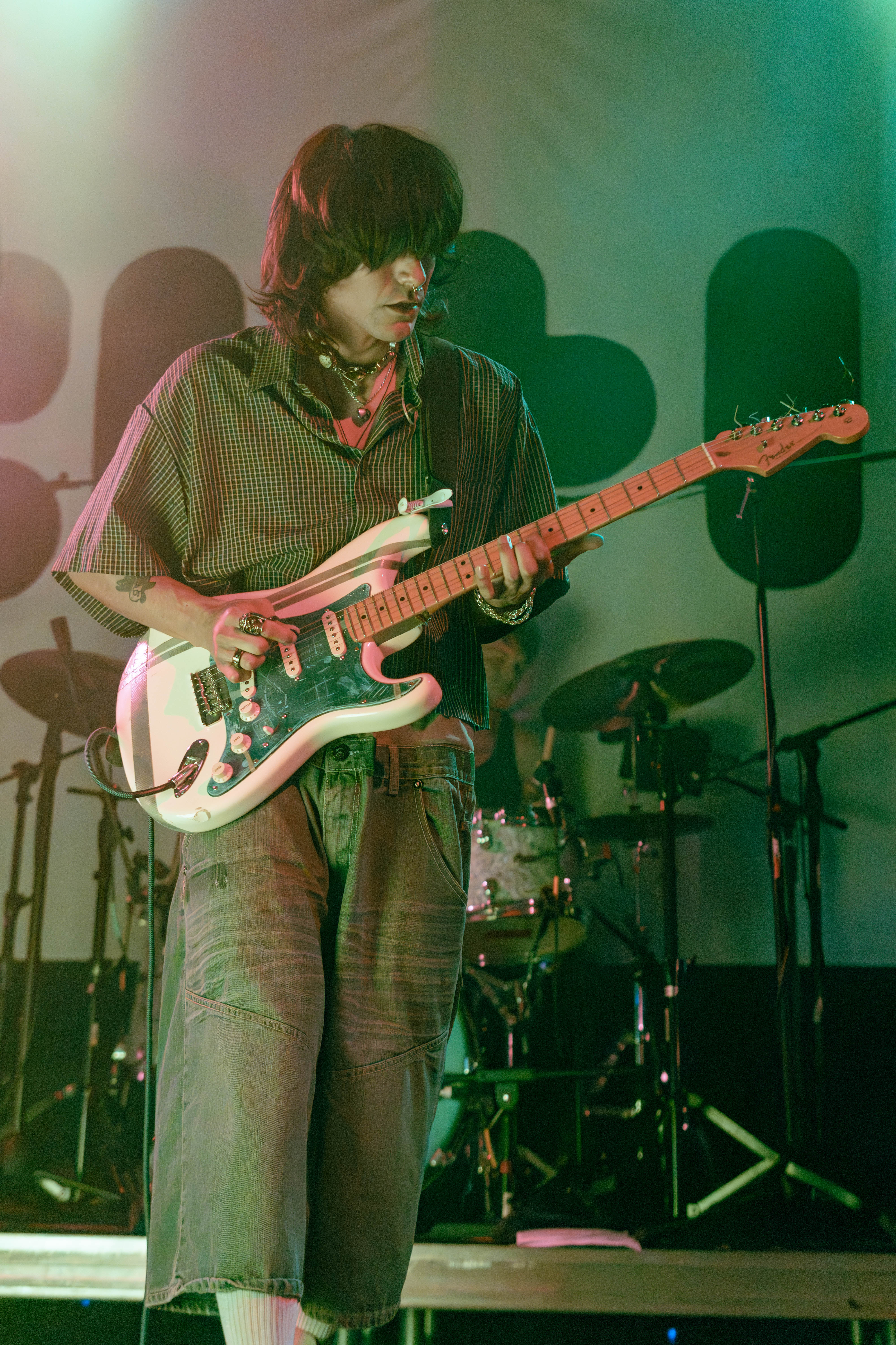 A musician playing guitar on stage in a vibrant, illuminated setting, wearing a checkered shirt and loose-fitting pants, with a focus on their intense expression.